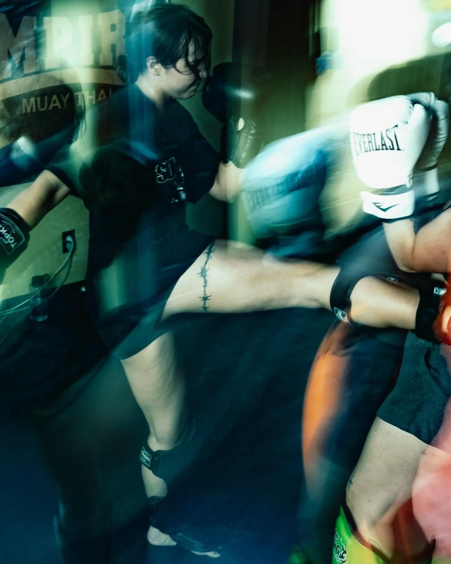 A woman practicing boxing with a trainer through a glass window in a gym. The woman is wearing boxing gloves and shorts, and the trainer is holding a mitt for her to punch.