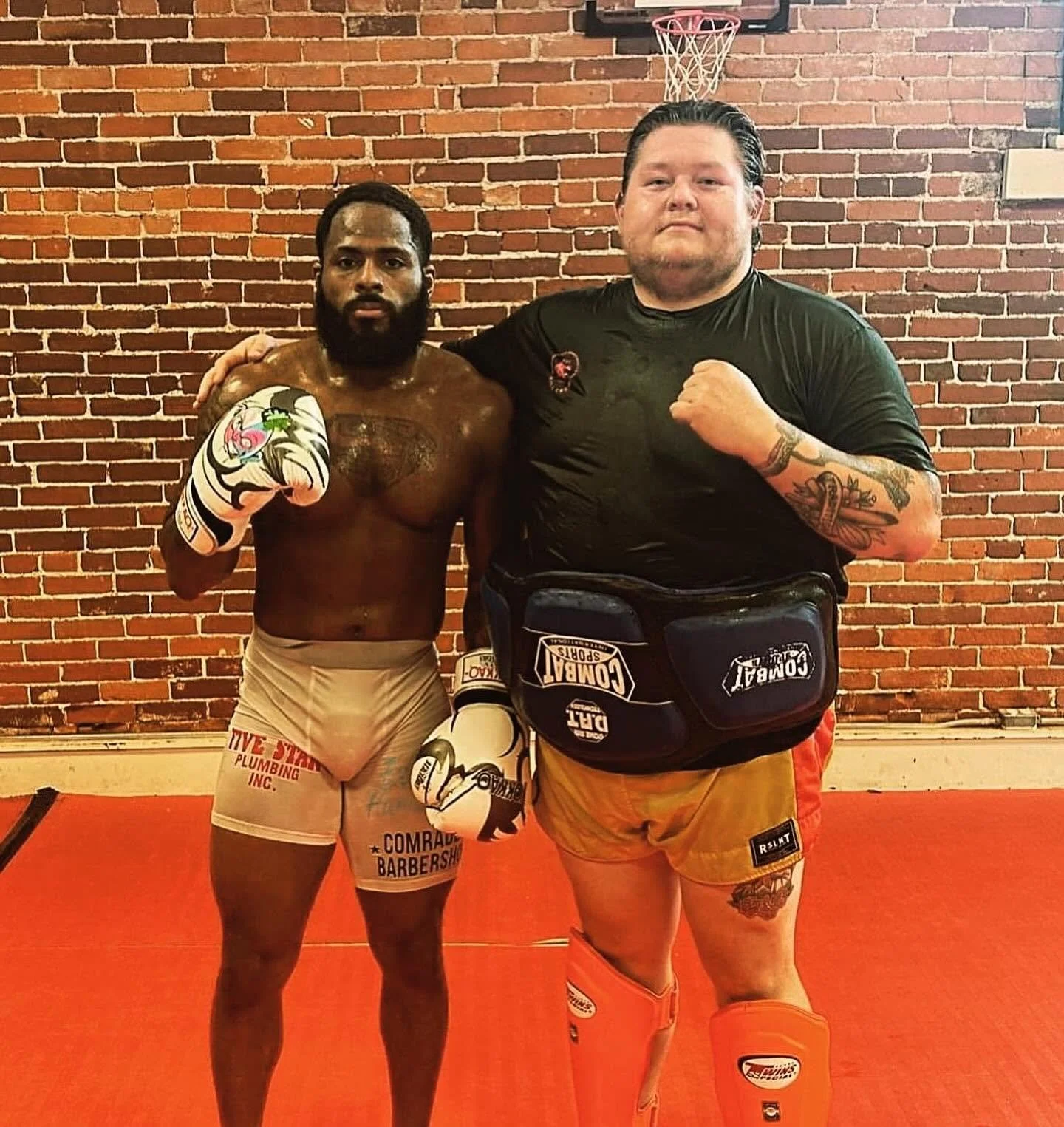 Two men in a gym with a brick wall background, one shirtless with boxing gloves, the other wearing a black t-shirt and shorts, standing with fists raised.