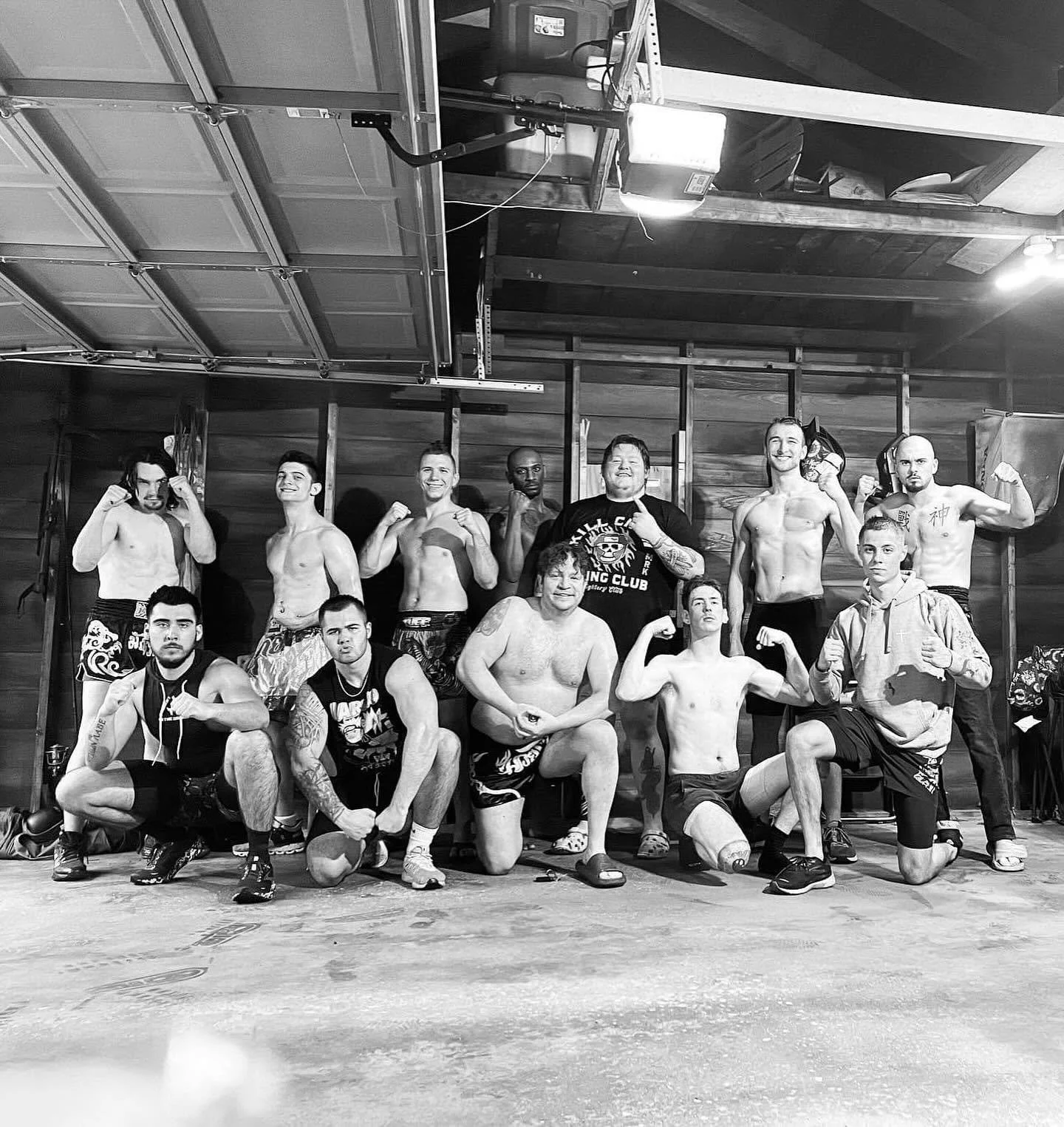 Group of men in a gym, some shirtless, posing with fists and flexed muscles, sitting and standing in front of a wooden wall.