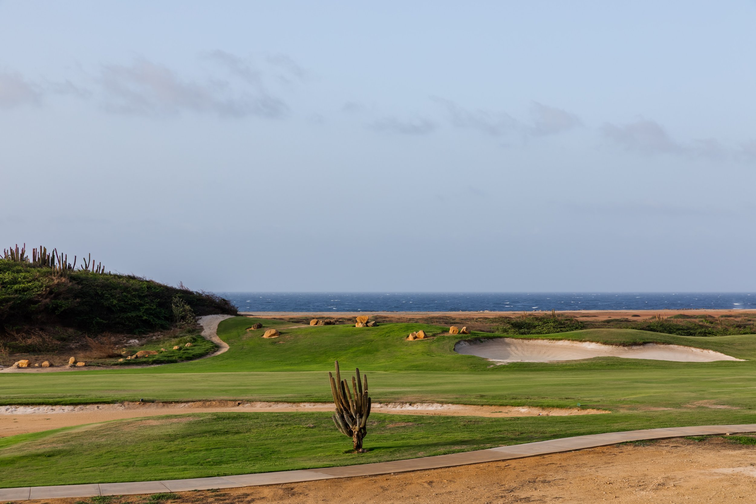 A coastal golf course with green fairways, sand bunkers, and a view of the ocean under a cloudy sky.