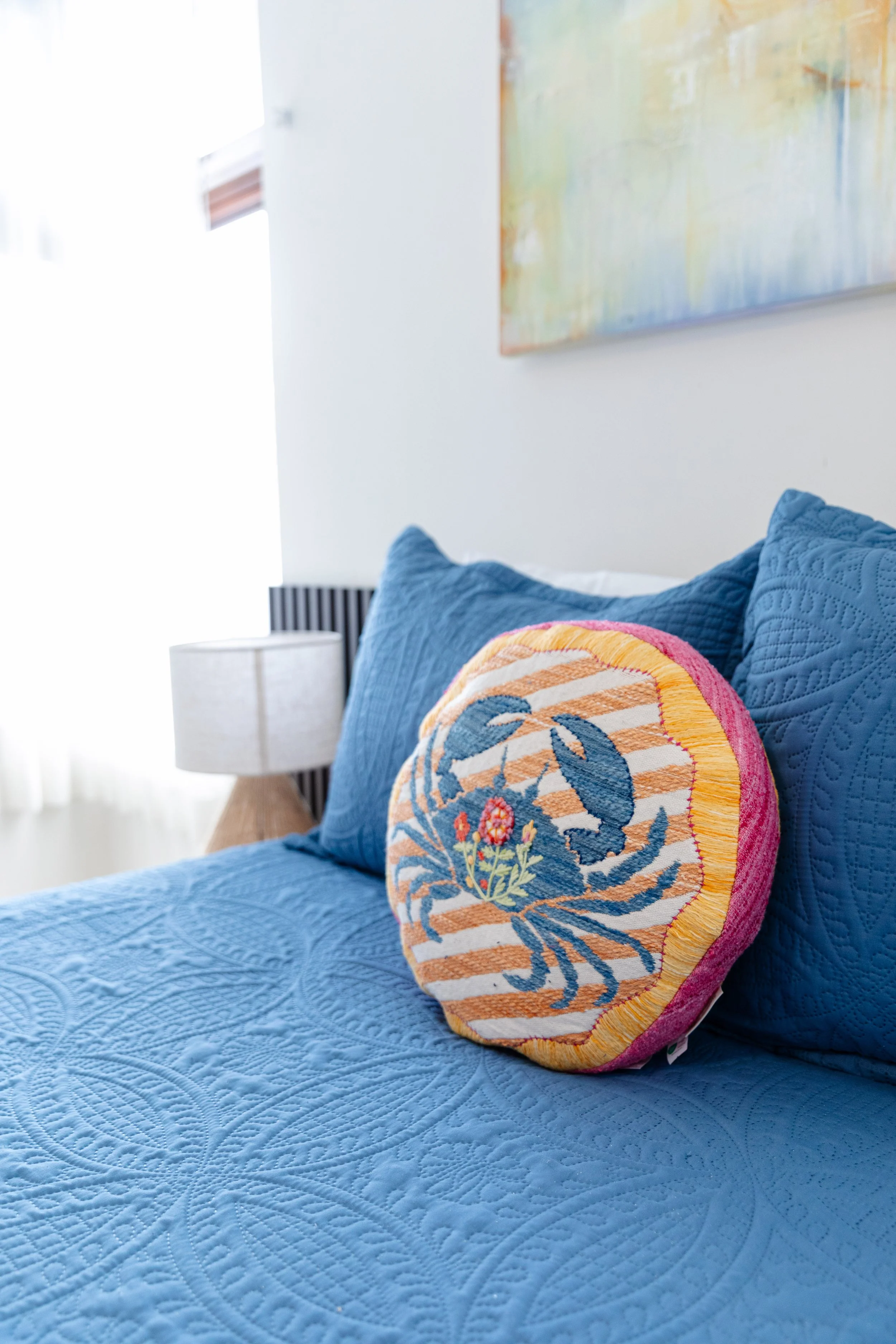 Close-up of a decorative pillow with an embroidered floral and butterfly design on a bed with a blue quilted bedspread, with a nightstand and a lamp in the background