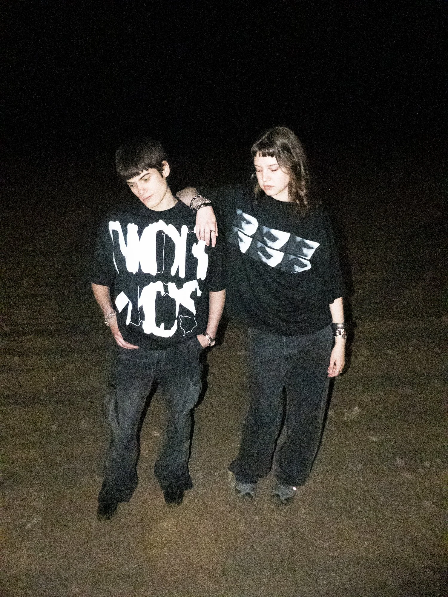 Two young women standing outdoors at night, with one woman resting her arm on the other's shoulder, both wearing black graphic T-shirts and dark pants.