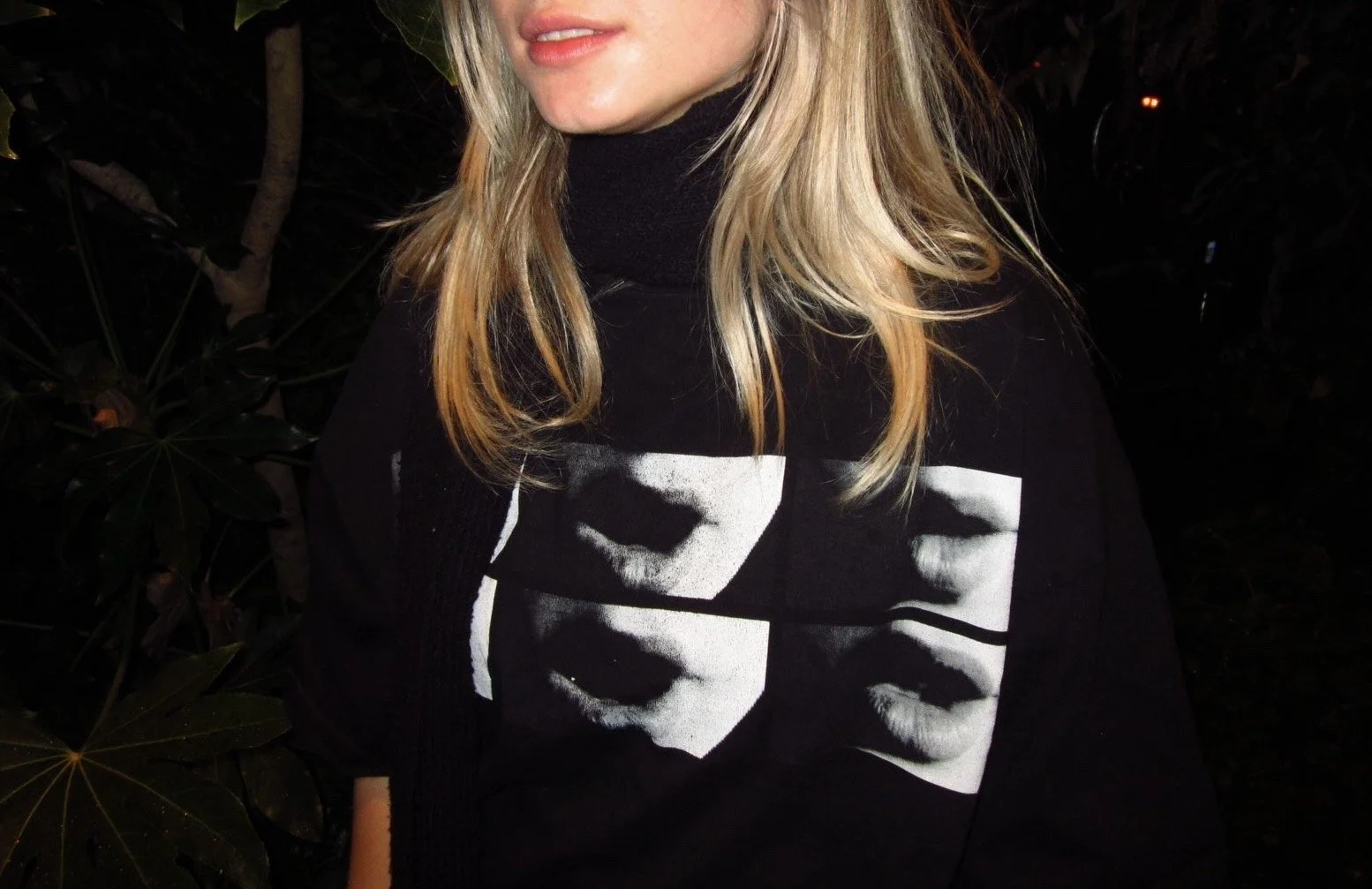 A woman with blonde hair wearing a black sweater with white graphic prints of lips and faces, standing outdoors at night.
