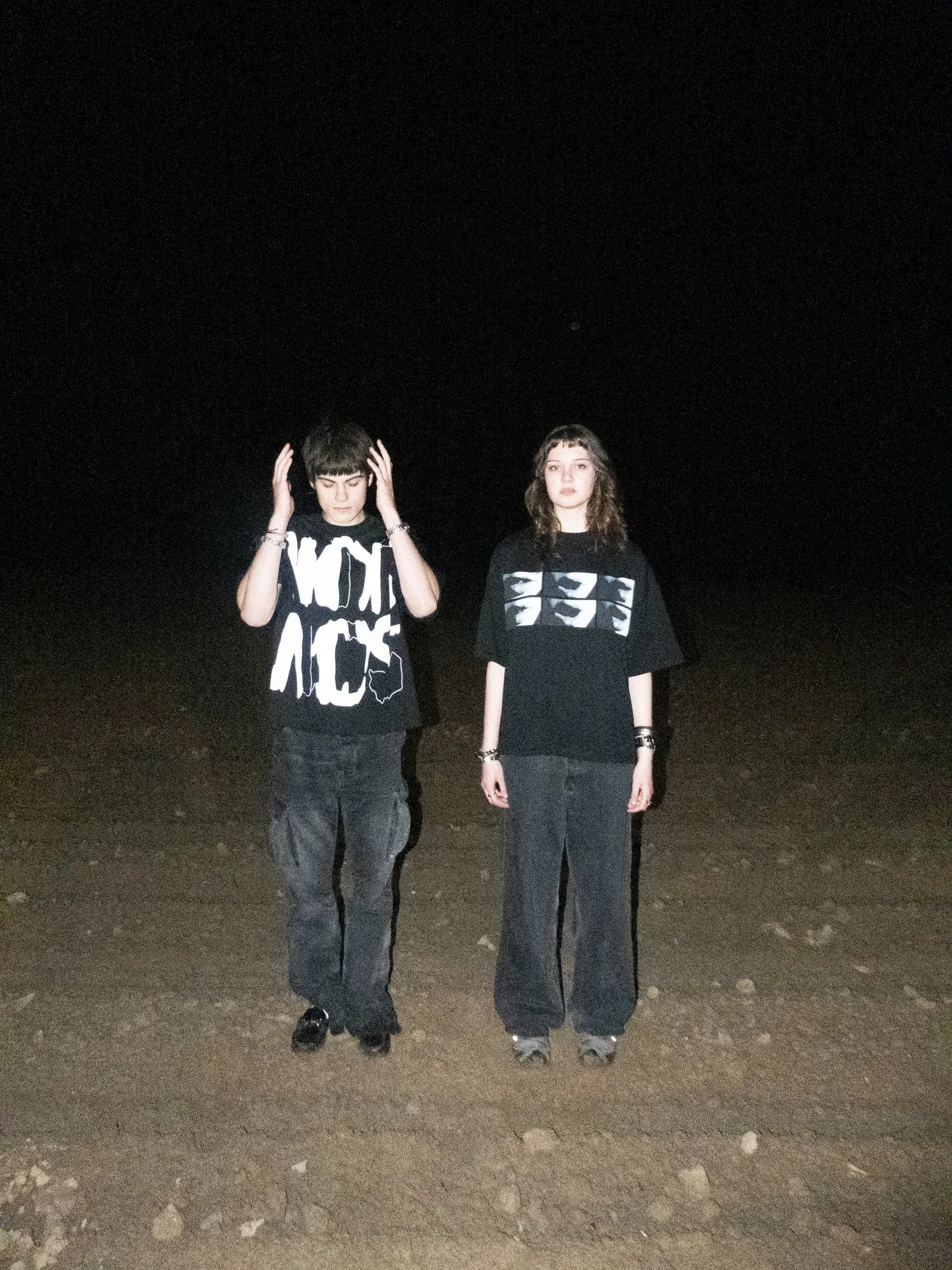 Two young women standing outdoors at night on a dirt ground, both wearing black oversized graphic T-shirts and dark pants. The woman on the left has short dark hair, is holding her hands to her head, and has a serious expression. The woman on the right has shoulder-length dark hair with bangs, has a neutral expression, and is looking at the camera.