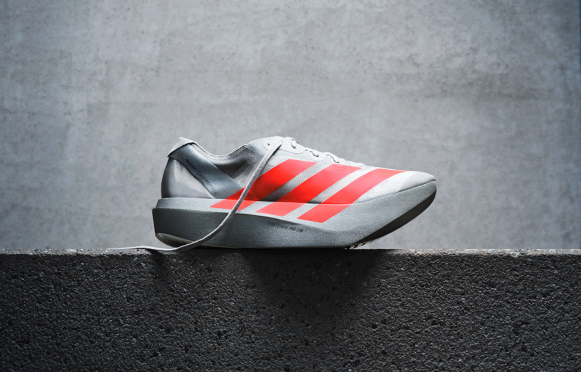 White running shoe with red stripes resting on a concrete surface against a gray wall background.