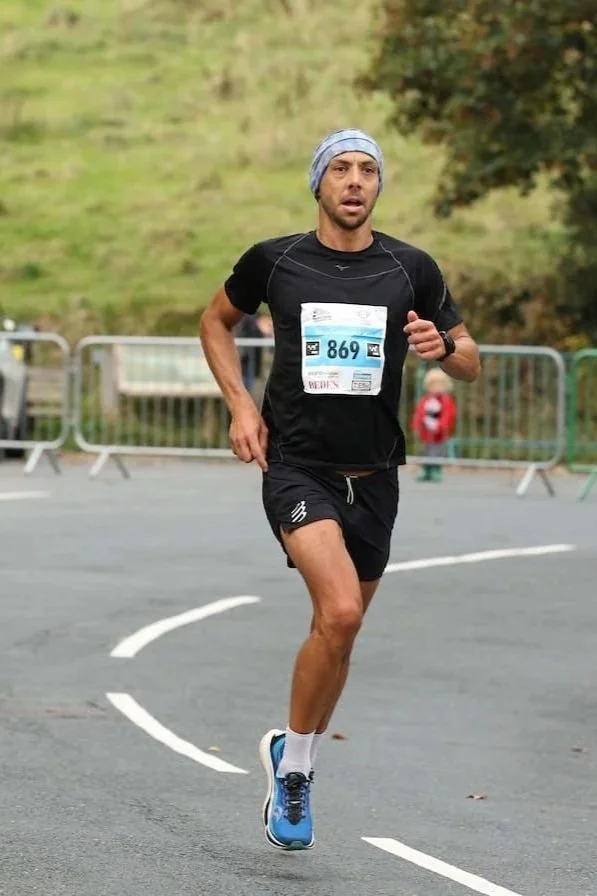 A male runner with a race bib numbered 860, wearing a black shirt, black shorts, blue running shoes, and a light-colored head covering, running outdoors on a paved road.