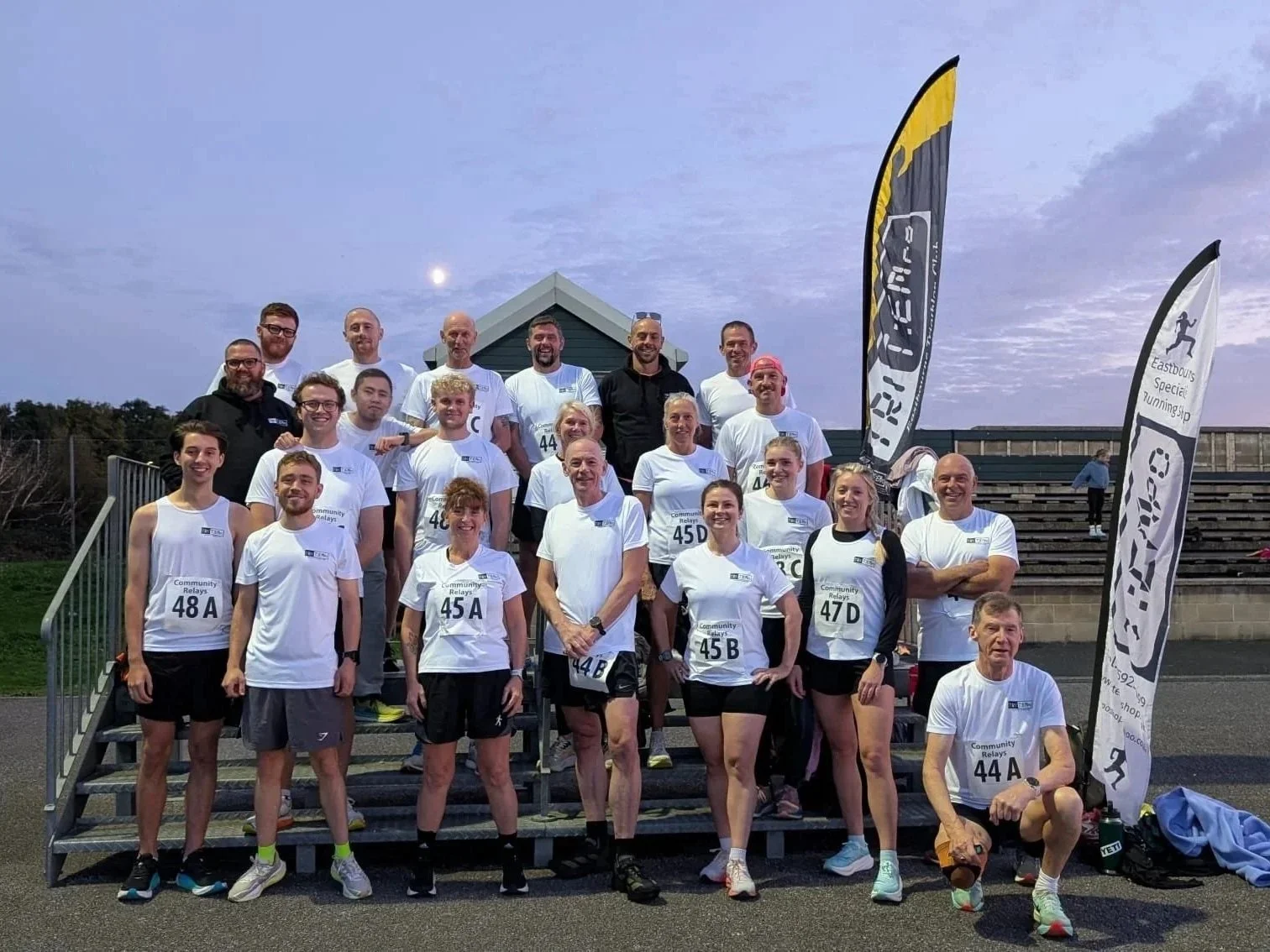 Group of people in athletic attire participating in a community relay race, standing on a tiered podium with flags and a wooden building in the background during evening time.