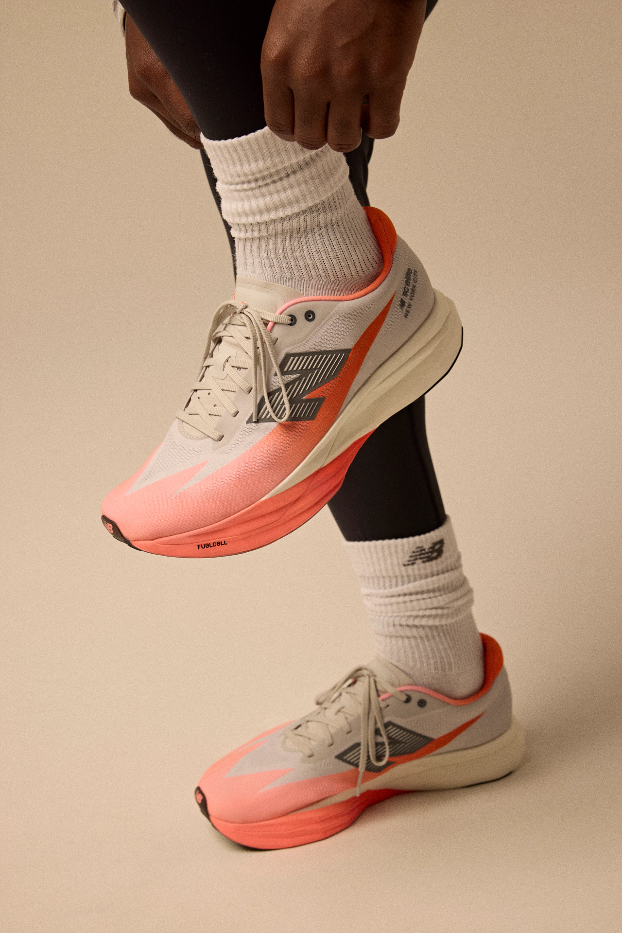 A person wearing white New Balance sneakers with orange accents, white socks, and black leggings is tying the shoelaces, standing on a beige background.
