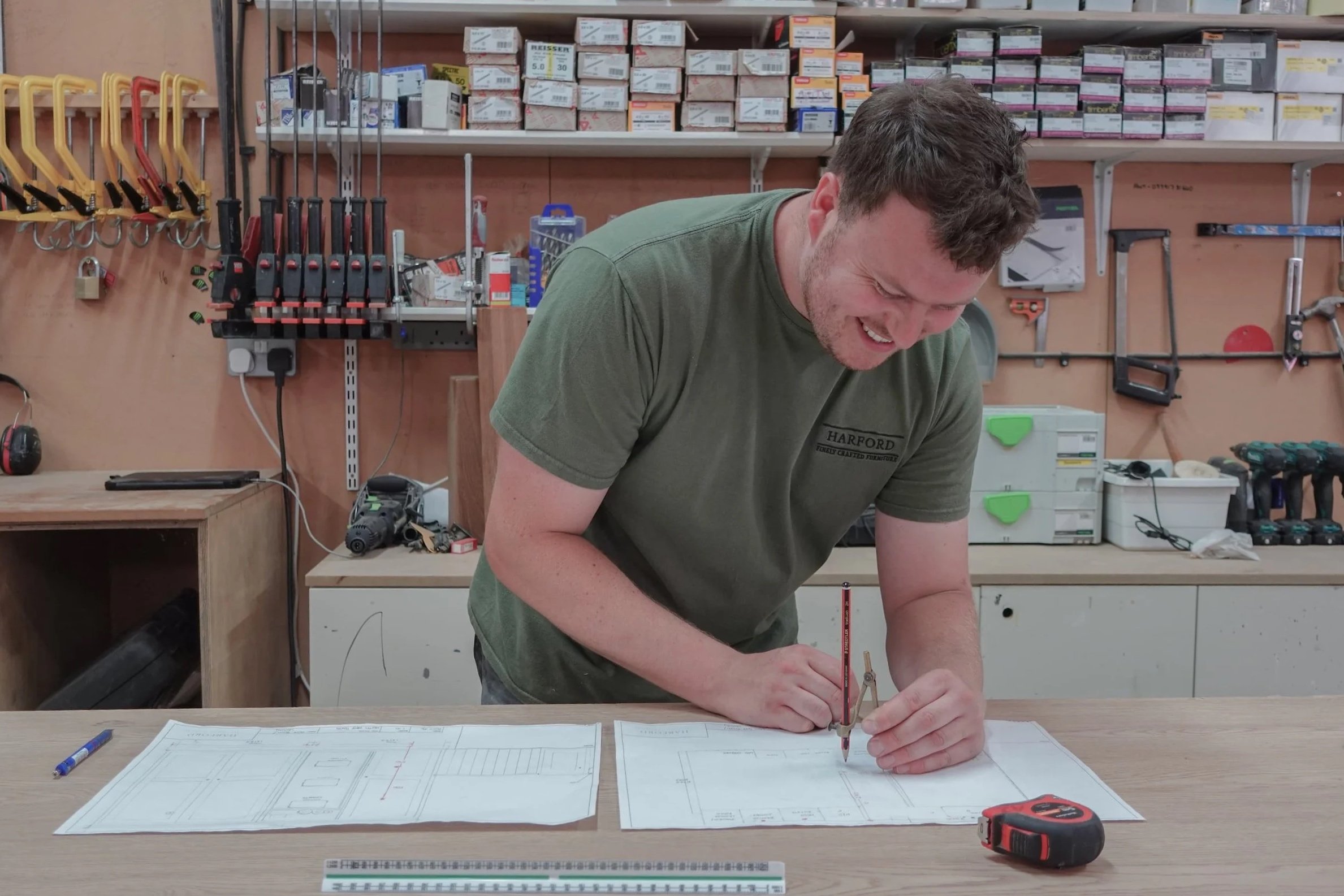 A man wearing a green t-shirt, holding a red pencil, drawing plans for a bespoke kitchen area. He has a ruler and tape measure on the desk and is in a workshop setting.
