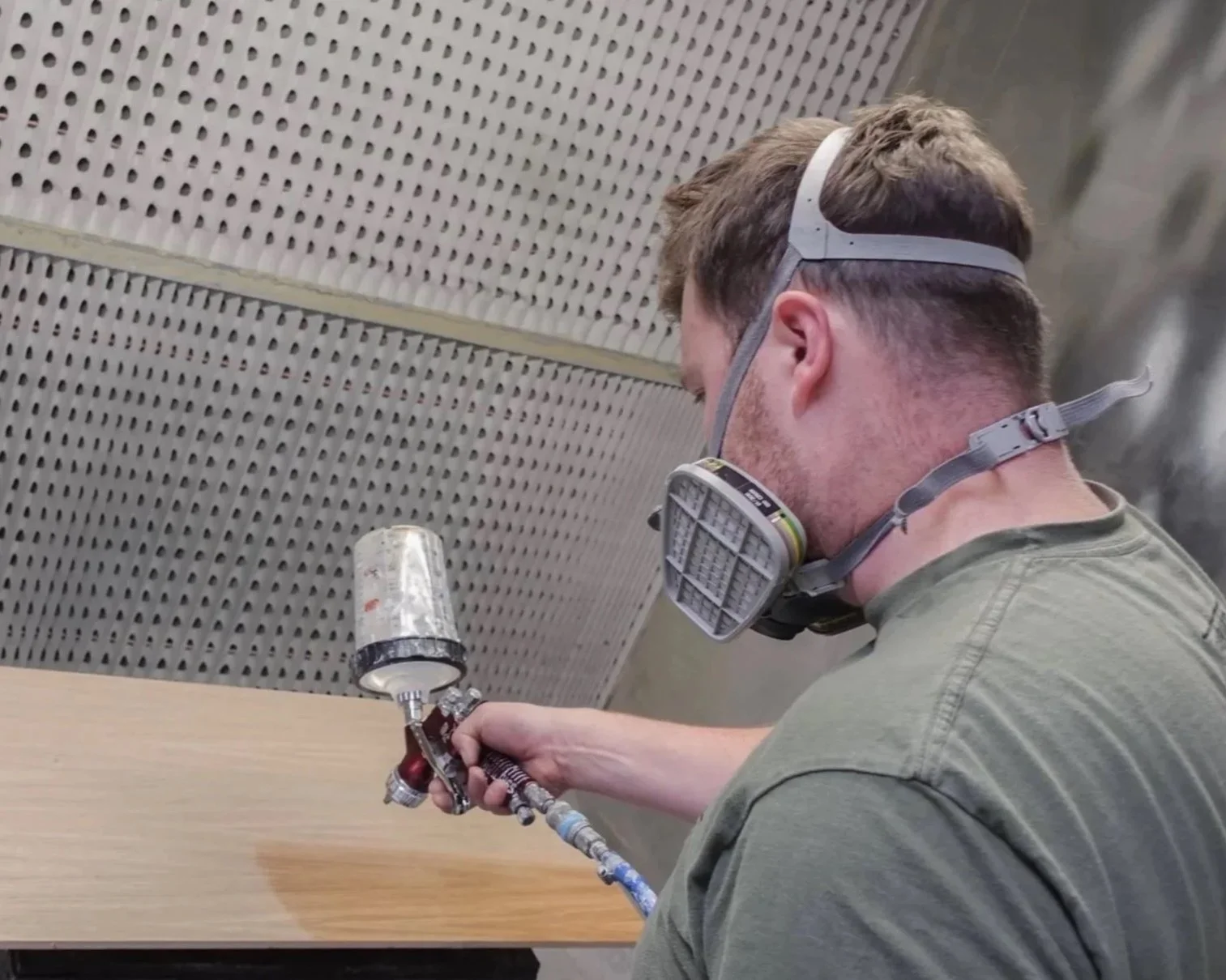 A man in a green t-shirt in a spray booth, spraying a piece of wood with a gloss.