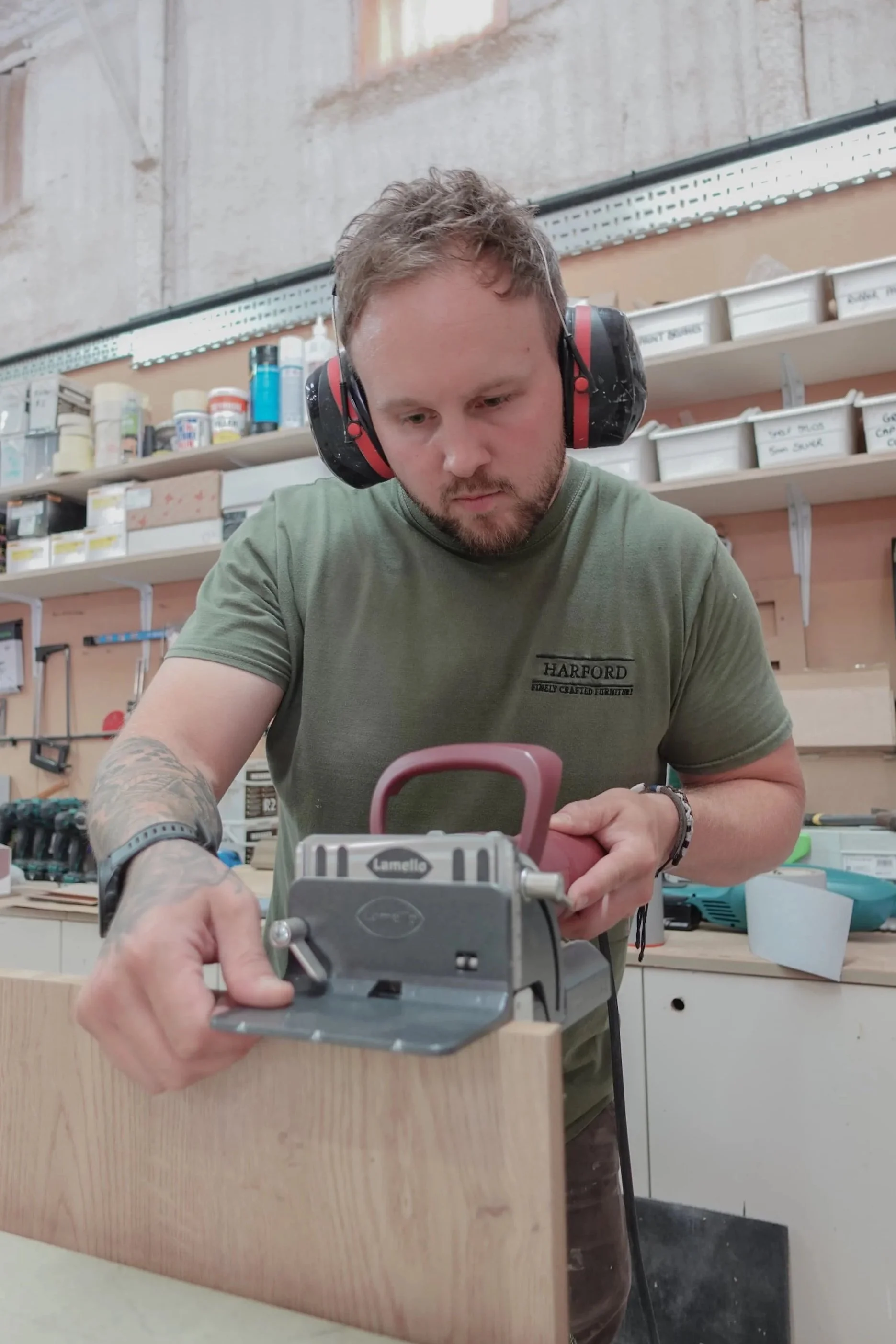 A man wearing a green t-shirt wearing ear defenders, using an electric hole machine to punch out hole in a piece of wood. In a workshop setting.