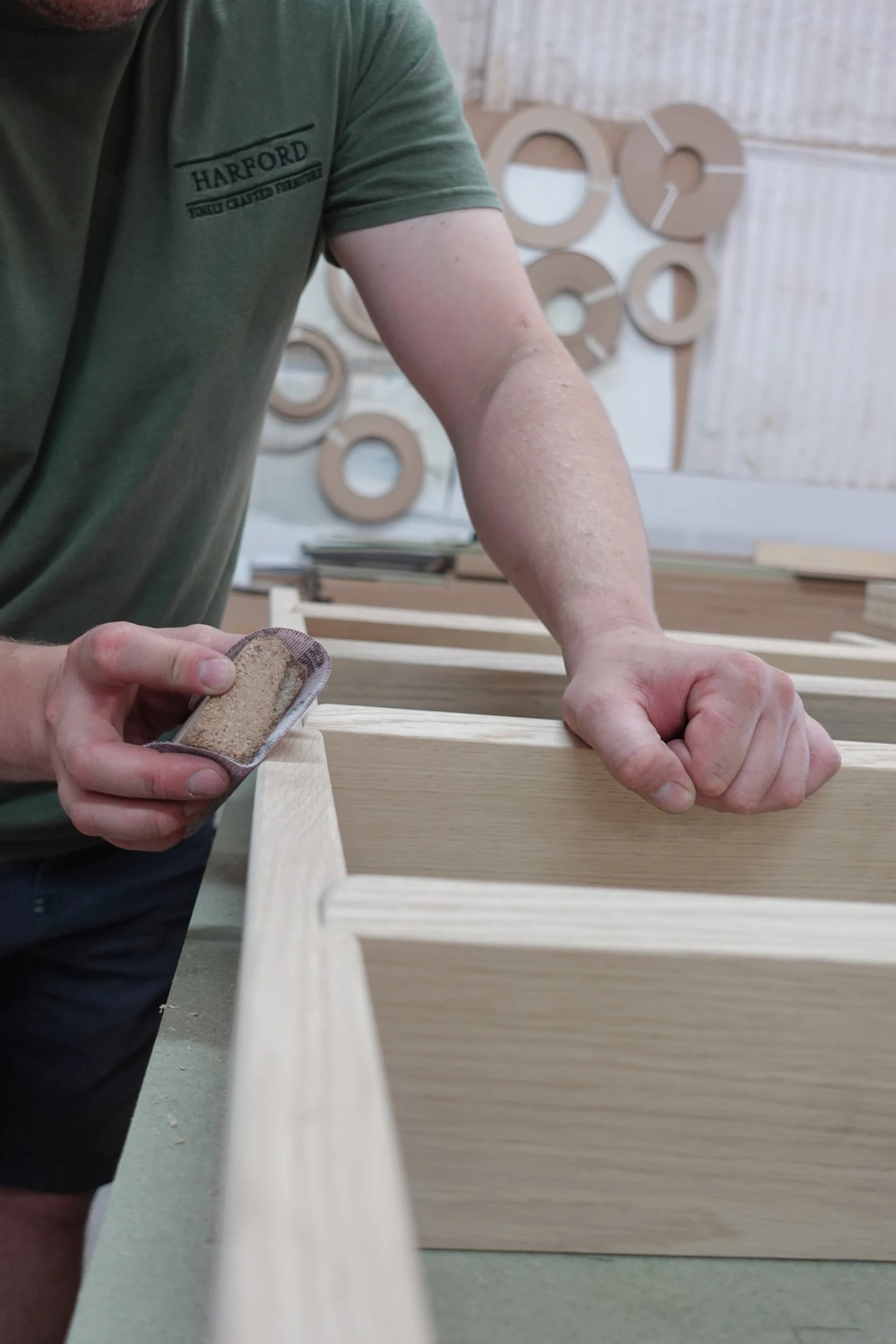 A close up on a man wearing a green t-shirt, hand sanding a handmade wooden ladder in a workshop setting.