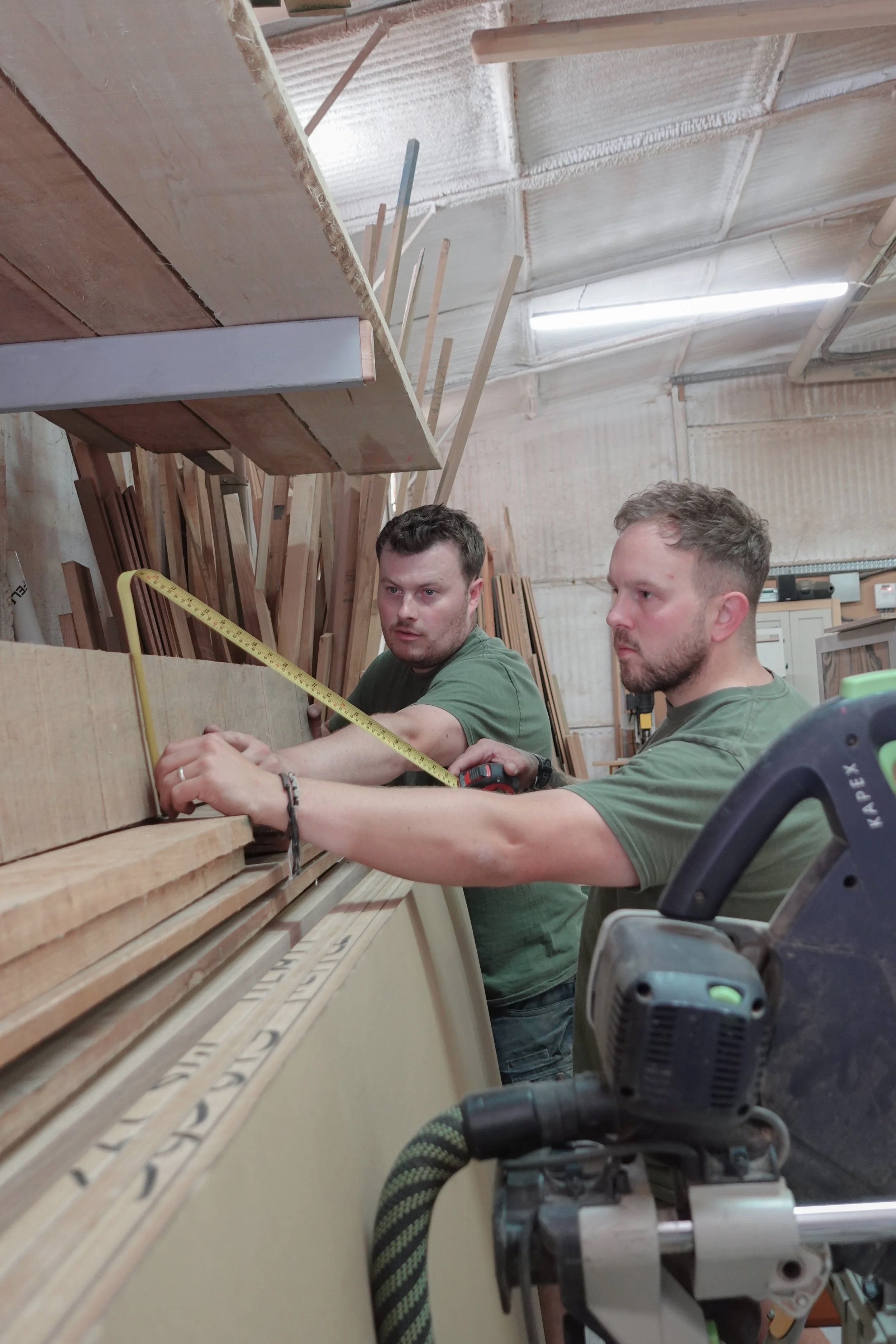 Two men wearing green t-shirts measuring up some wood in a workshop setting.