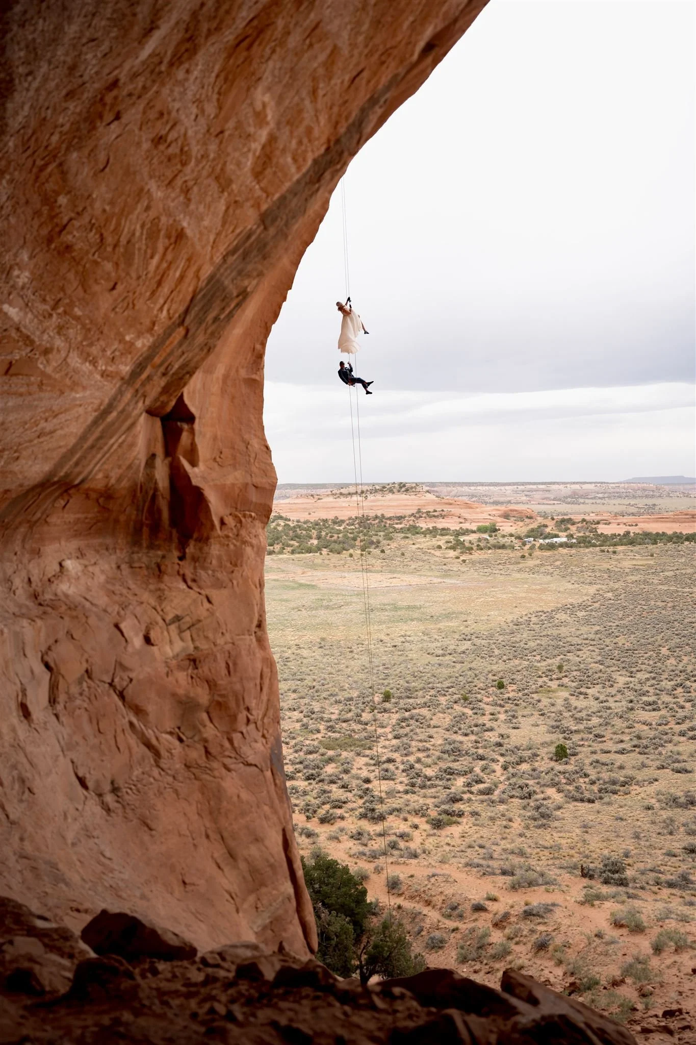 A person is hanging from a cliff with a swing, in a desert landscape with shrubs and distant mesas.