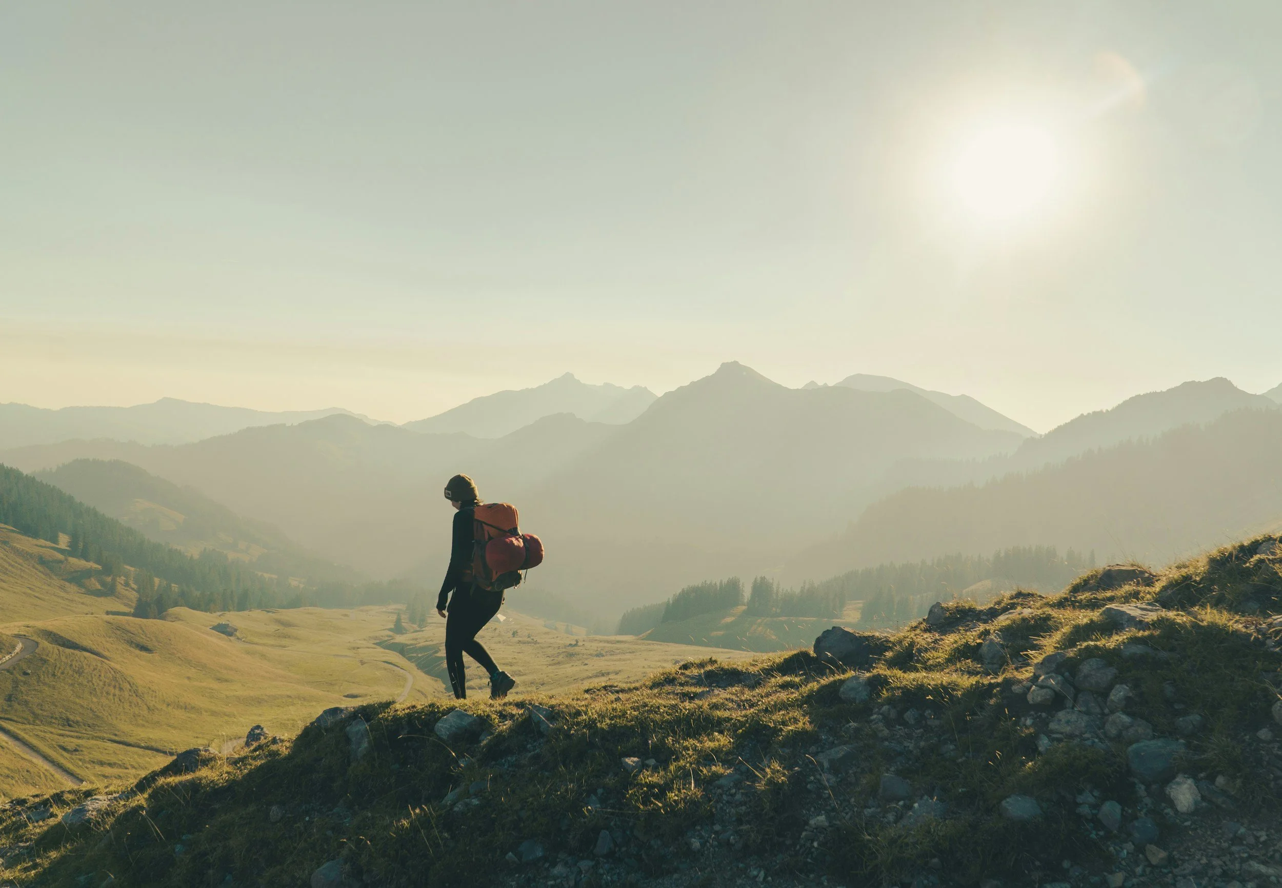 A person hiking in a mountainous landscape during the daytime, with rolling green hills, rugged mountain peaks in the background, and a hazy sky with the sun shining.