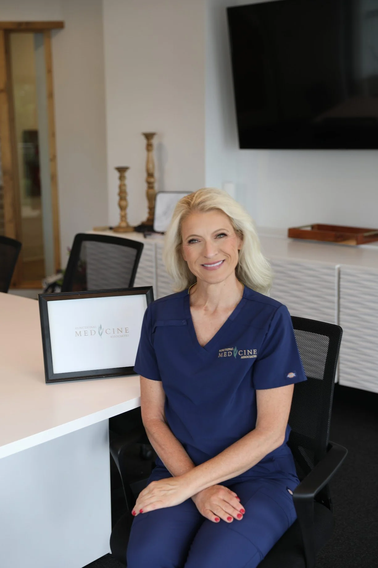 A blonde woman wearing navy medical scrubs sitting in a modern office environment. She is smiling and sitting with her hands on her lap. There is a framed sign on the desk that reads "Functional Med Care Associates". The background includes a monitor, decorative candlesticks, and a white textured cabinet.