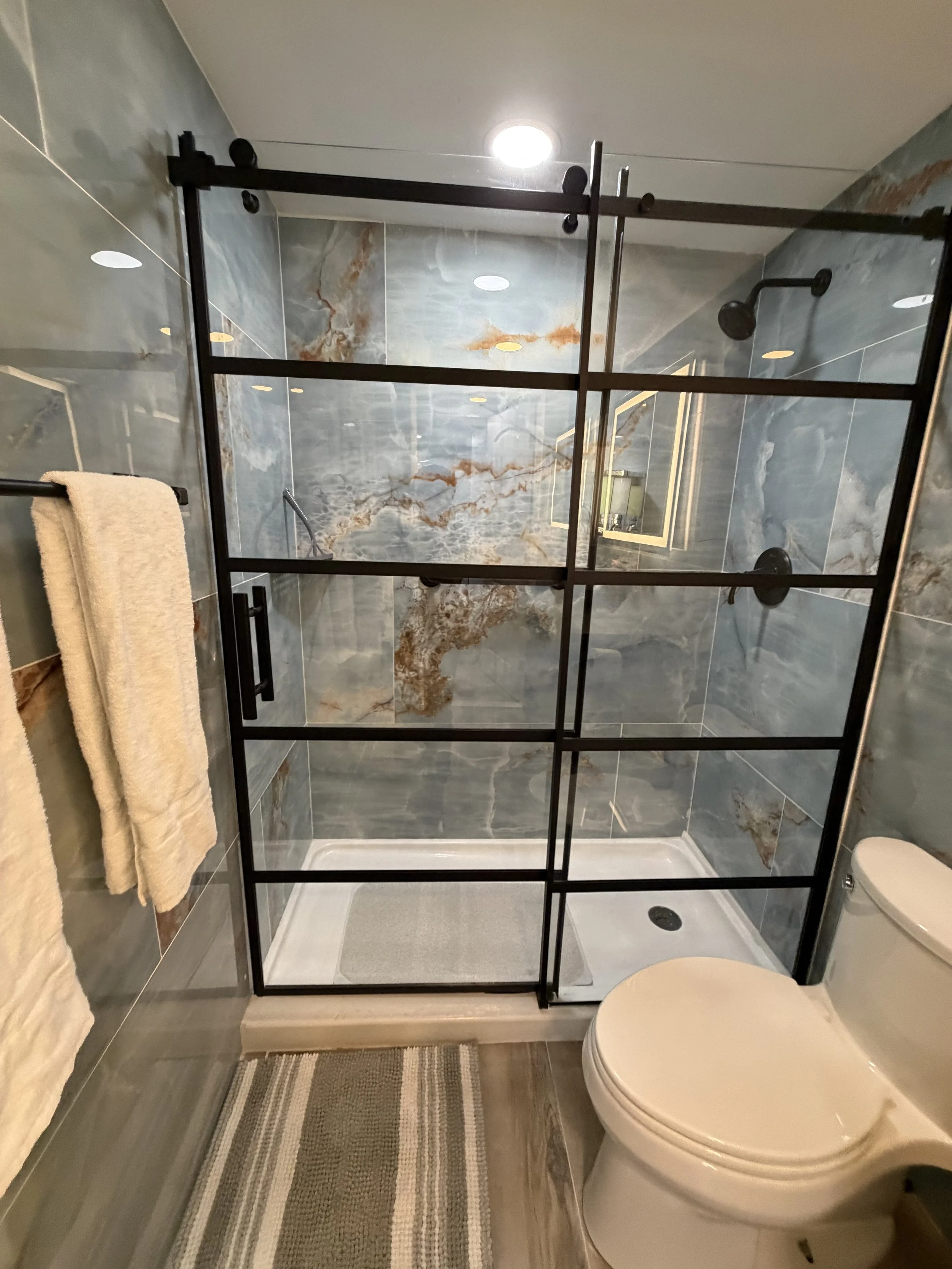 Bathroom with a walk-in shower enclosed by a black metal frame, marble walls, a towel on a towel rack, a toilet, and a striped rug on the floor.