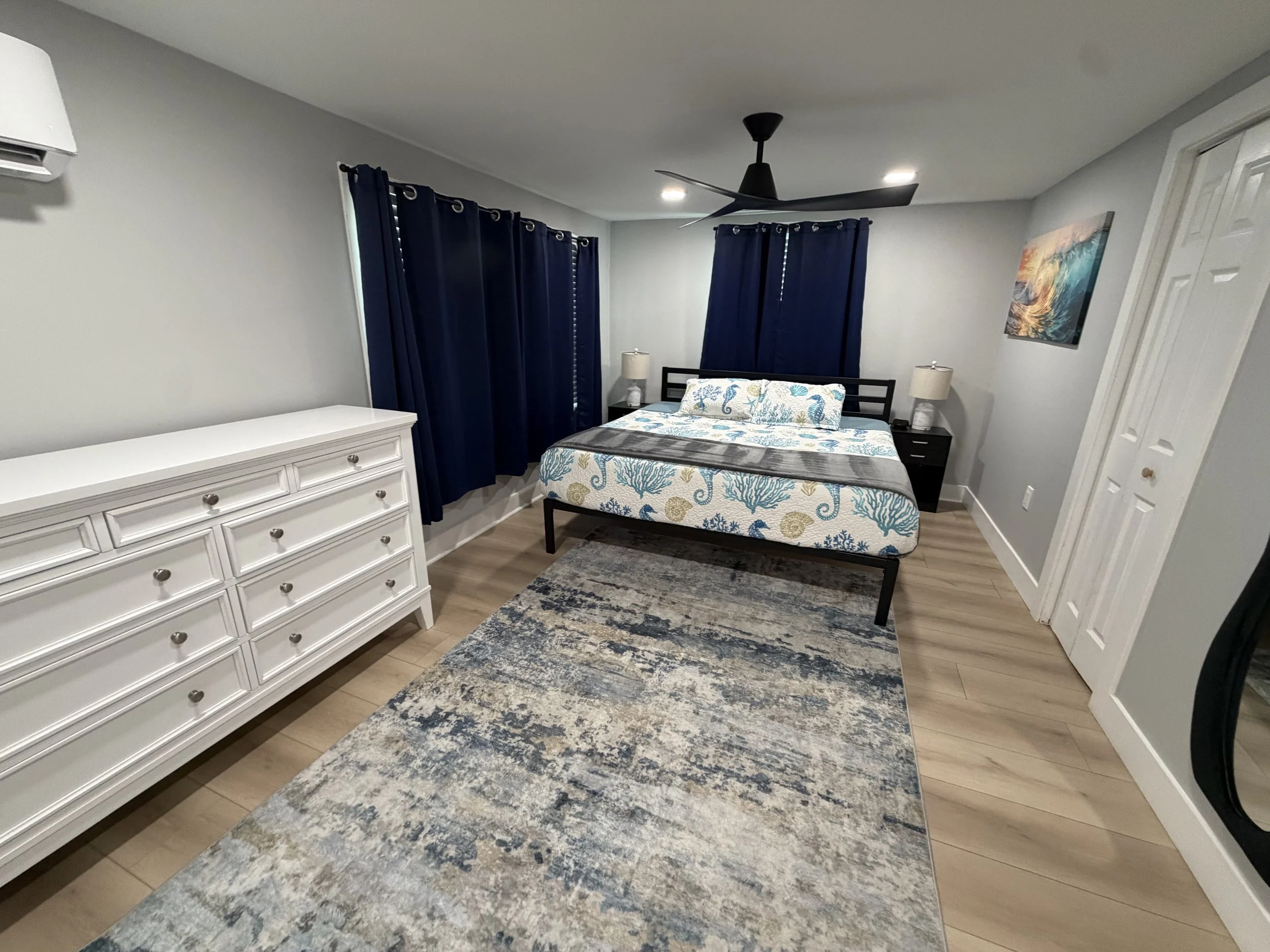 A bedroom with a queen-size bed, white dresser, dark blue curtains, and a striped area rug.
