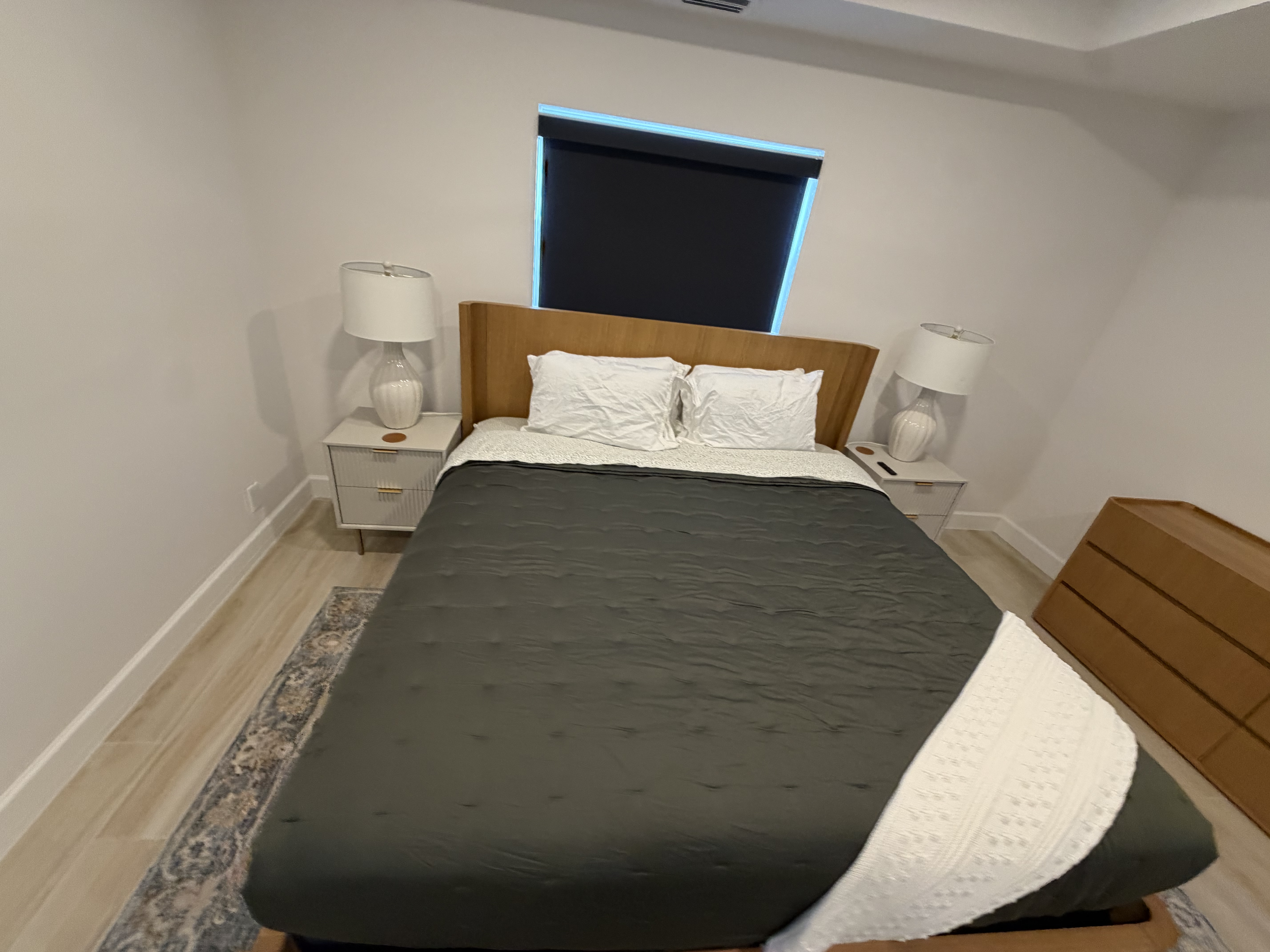 A bedroom with a wooden headboard, two white pillows, and a dark gray quilt. There are two nightstands with white lamps on either side of the bed. A window with closed blackout blinds is behind the headboard. The room has light-colored walls and light wood flooring.