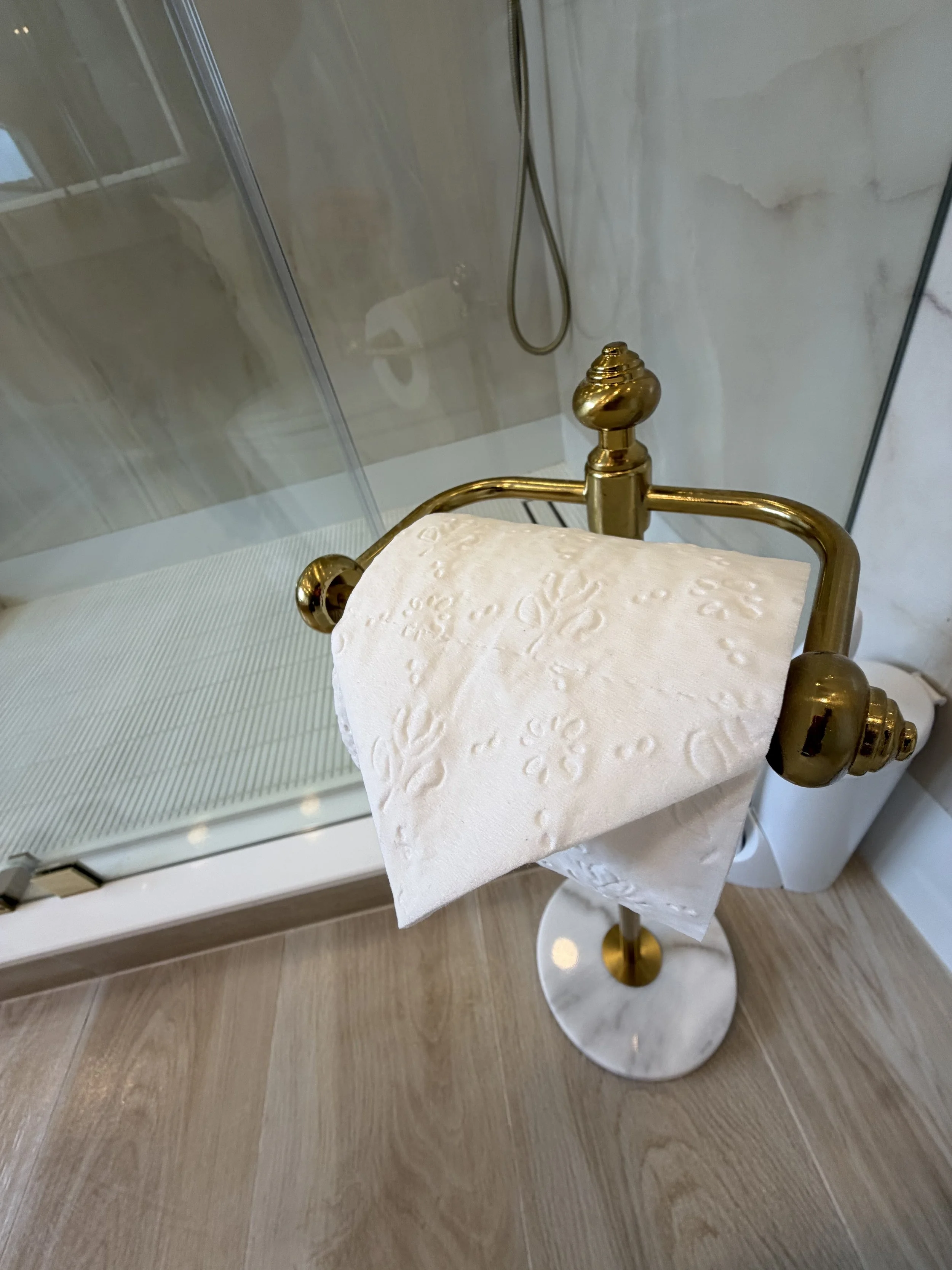 Bathroom with a gold toilet paper holder holding a roll of white toilet paper, beige marble bottom, and a shower with glass door and a hose.