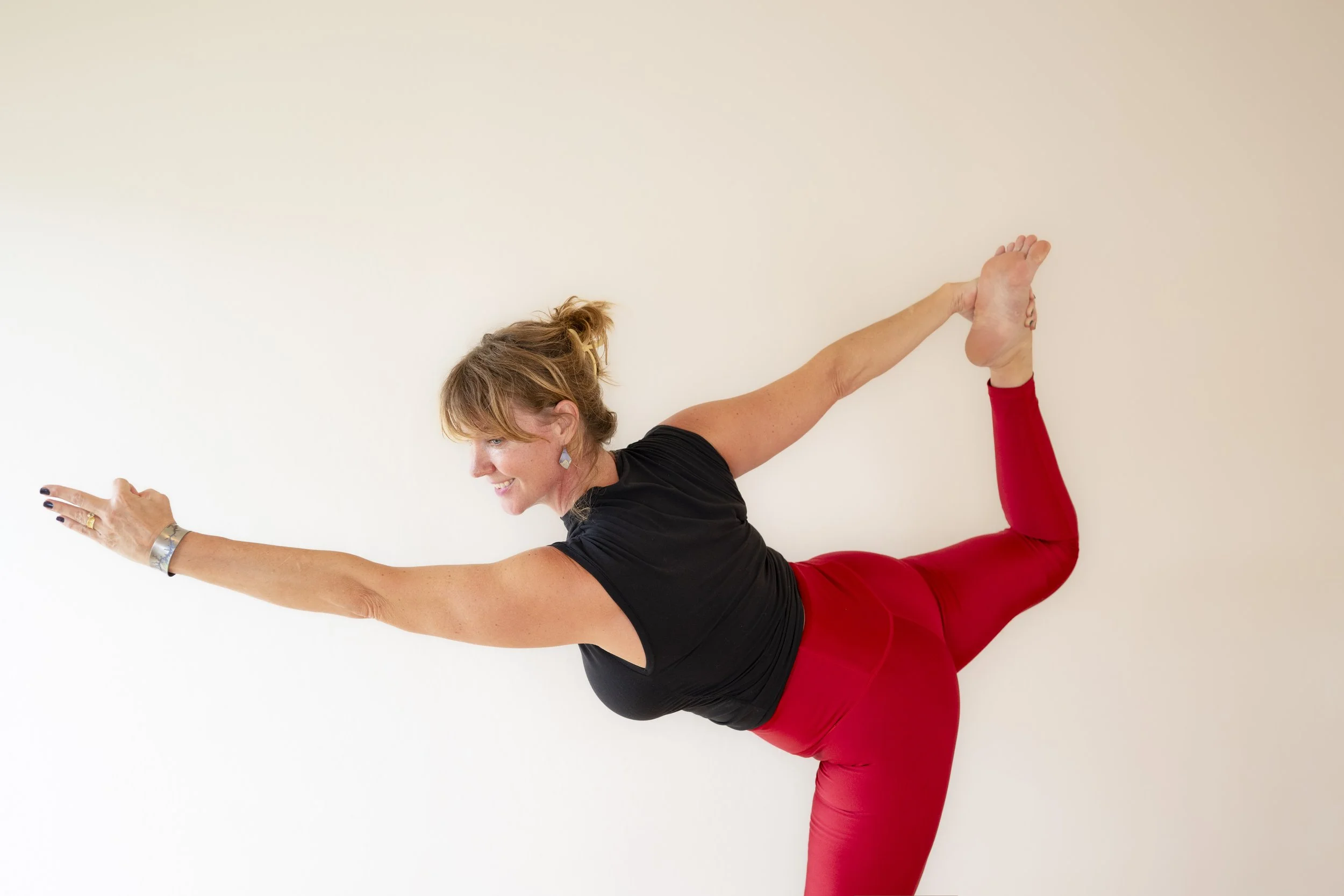A woman practicing yoga indoors, wearing red leggings and a black top, balancing on her left leg with her right leg bent behind her and her right foot held by her right hand, while extending her left arm forward.