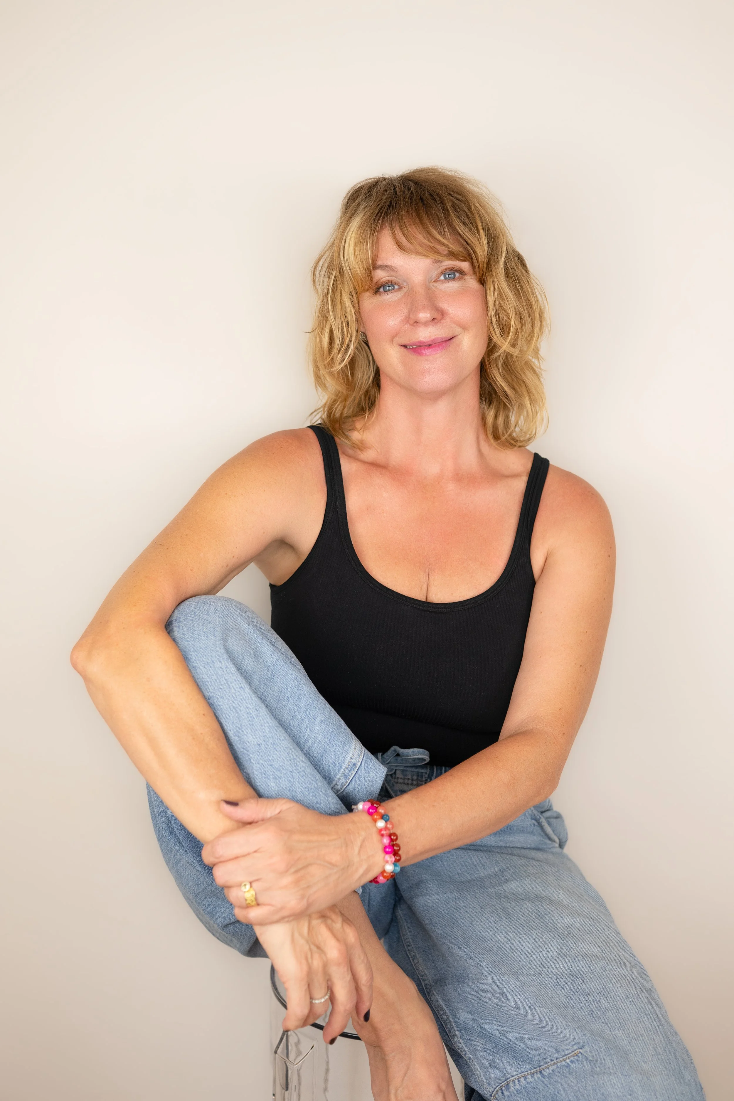 A woman with blonde hair, blue eyes, and fair skin, sitting casually against a plain light-colored wall, wearing a black tank top, blue jeans, and colorful beaded bracelets on her wrist, smiling gently at the camera.