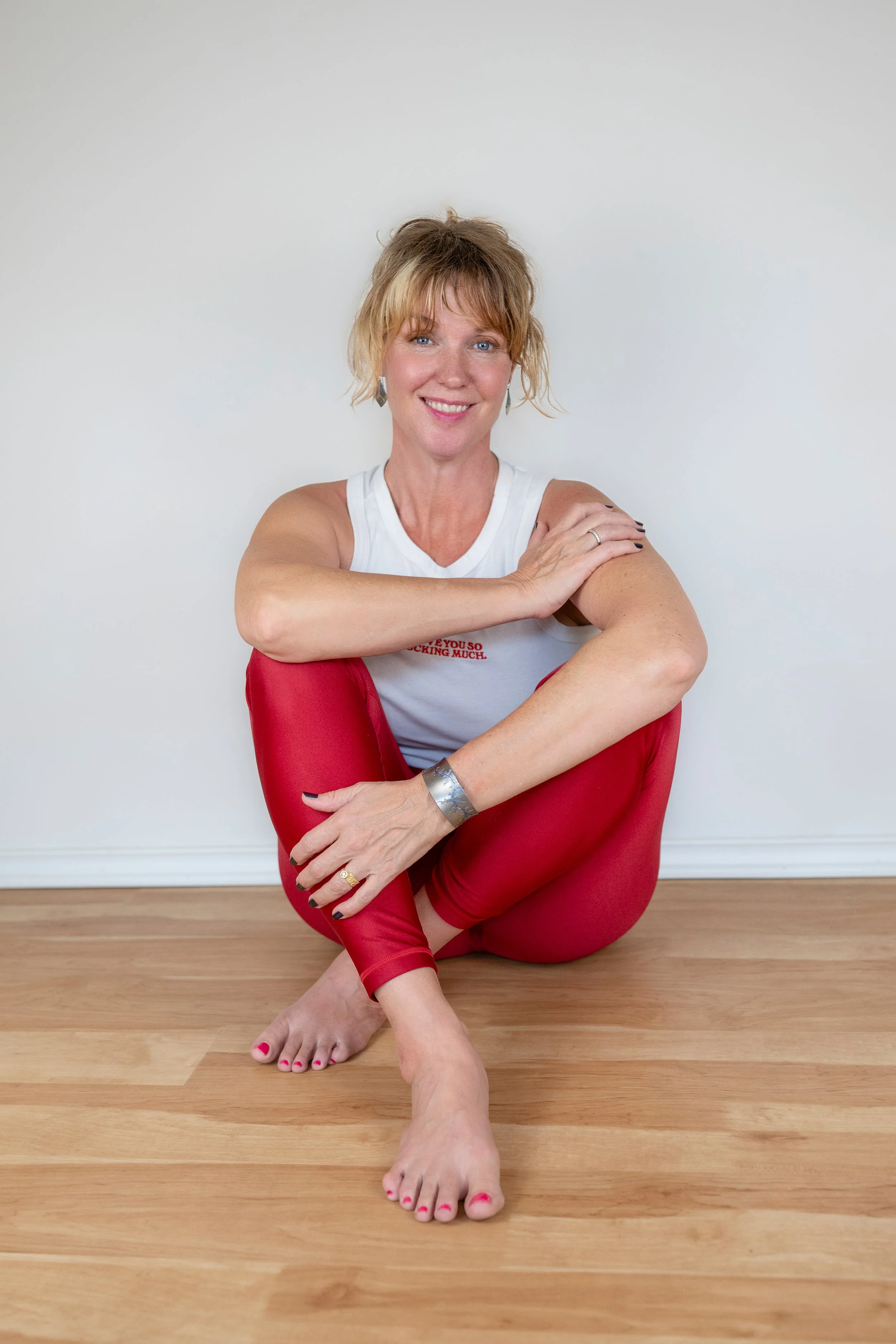 A woman with blonde hair and blue eyes, smiling, sitting on the wooden floor against a plain white wall. She is wearing a white sleeveless top with red text on it, red pants, and jewelry including earrings, a bracelet, and a ring. She is barefoot with pink painted toenails, and her arms are crossed with her right hand resting on her left shoulder.