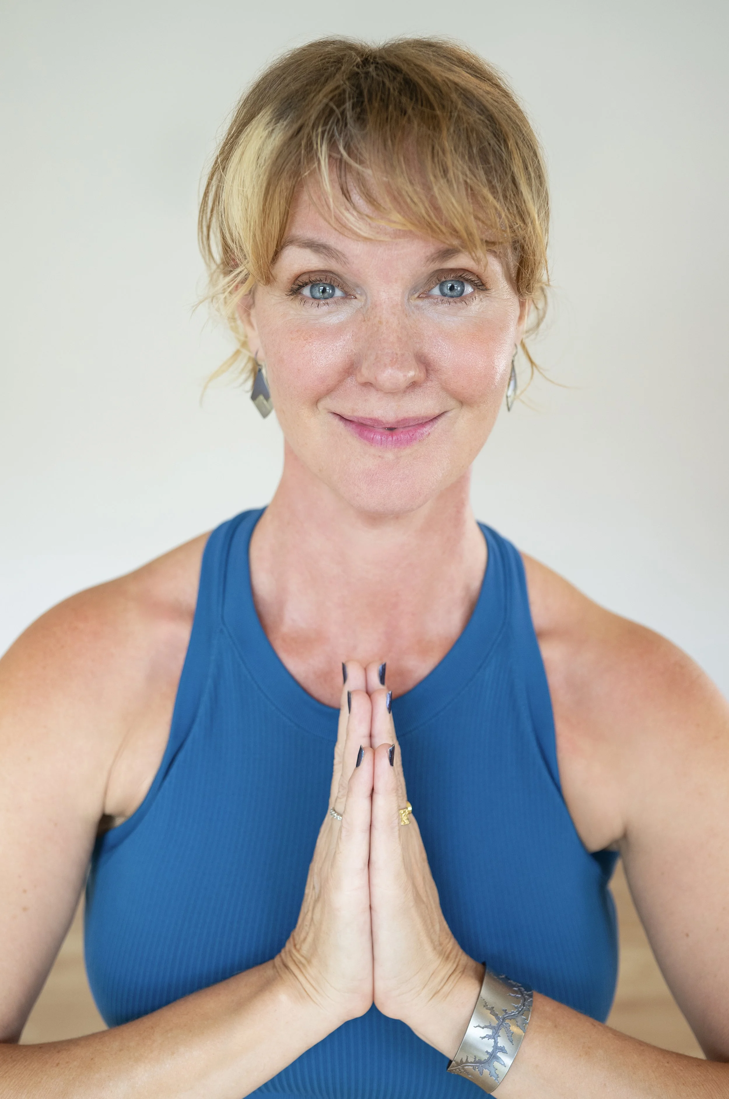 A woman with short red hair and blue eyes, wearing a blue tank top, is practicing yoga or meditation with her hands pressed together in front of her chest. She is smiling softly and wearing a silver bracelet and earrings.