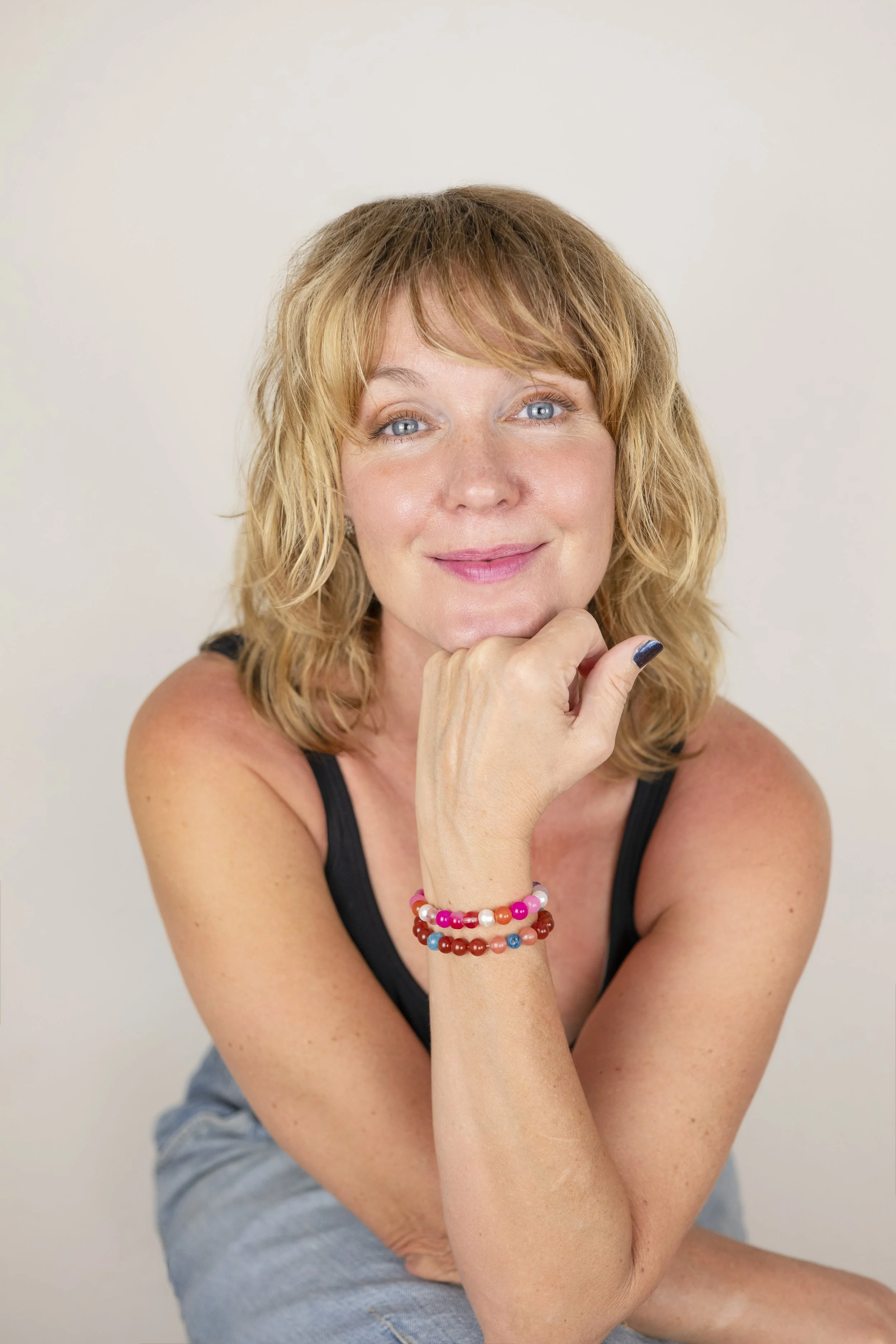 A woman with blonde hair and blue eyes smiling, resting her chin on her hand, wearing a black tank top and colorful beaded bracelets on her wrist.