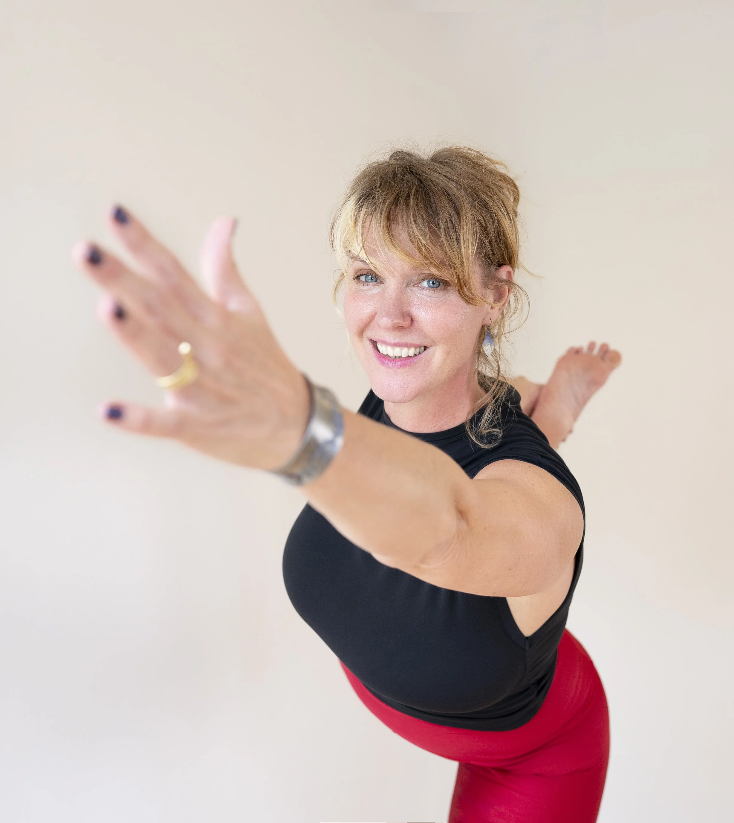 A woman with red hair and blue eyes looking at the camera, with her arm extended towards the lens, in a black top and red pants, standing against a plain white background.