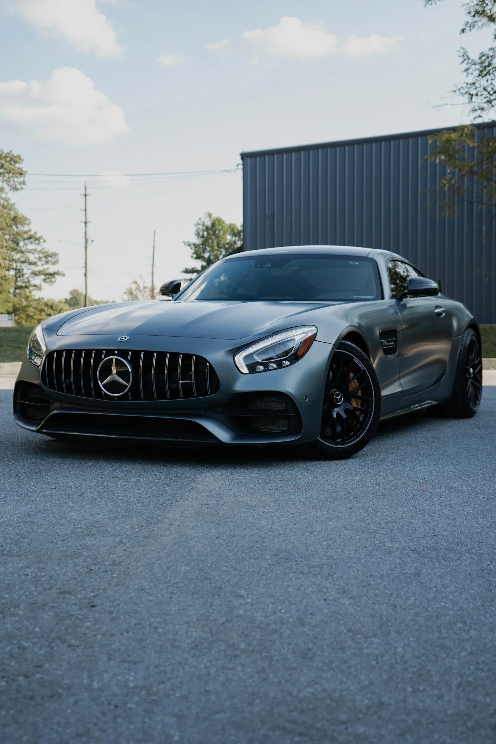 Gray Mercedes-Benz sports car parked outdoors near a blue building, trees, and power lines under a partly cloudy sky.