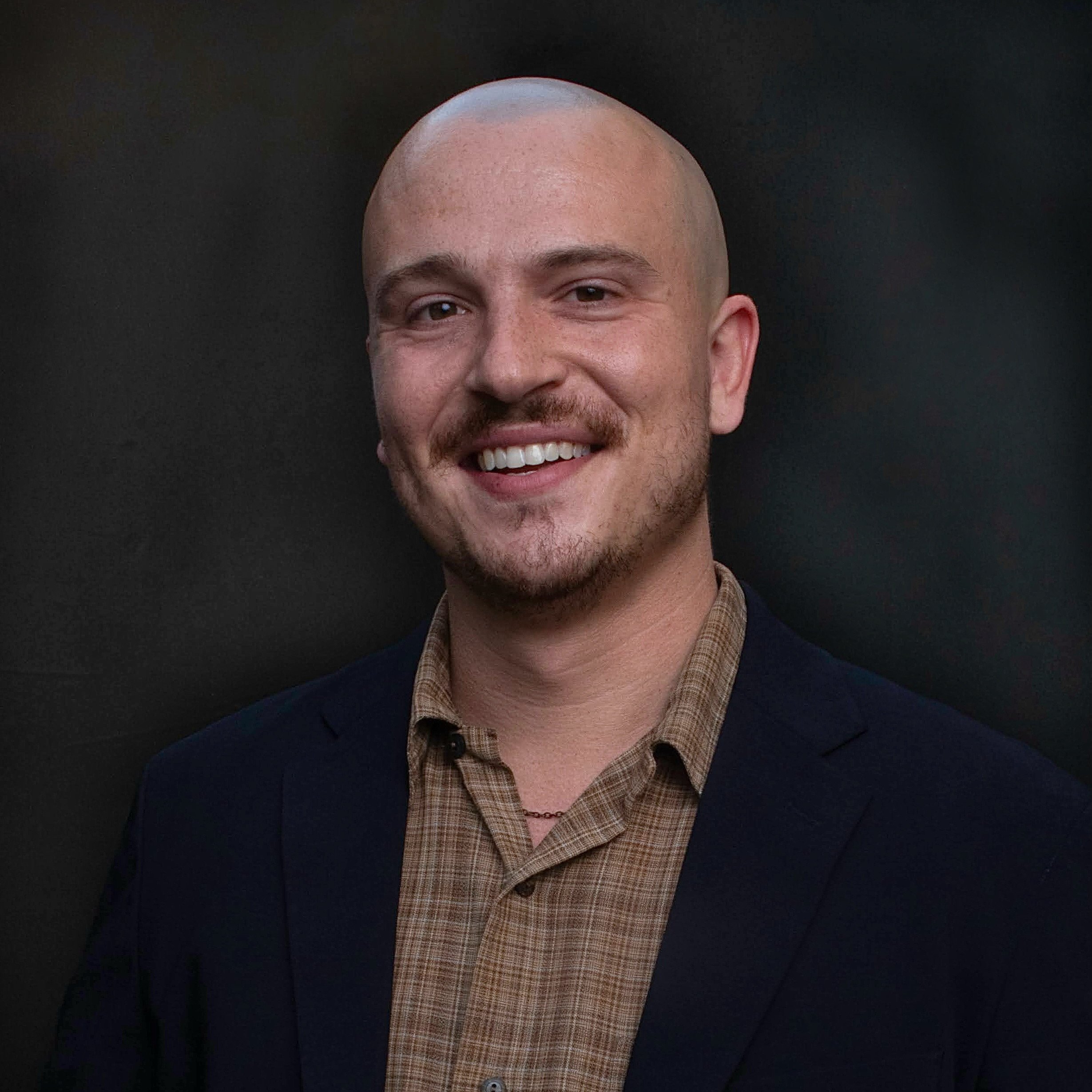 A smiling man with a shaved head, light skin, and facial hair, wearing a plaid shirt and a dark blazer, standing against a dark background.