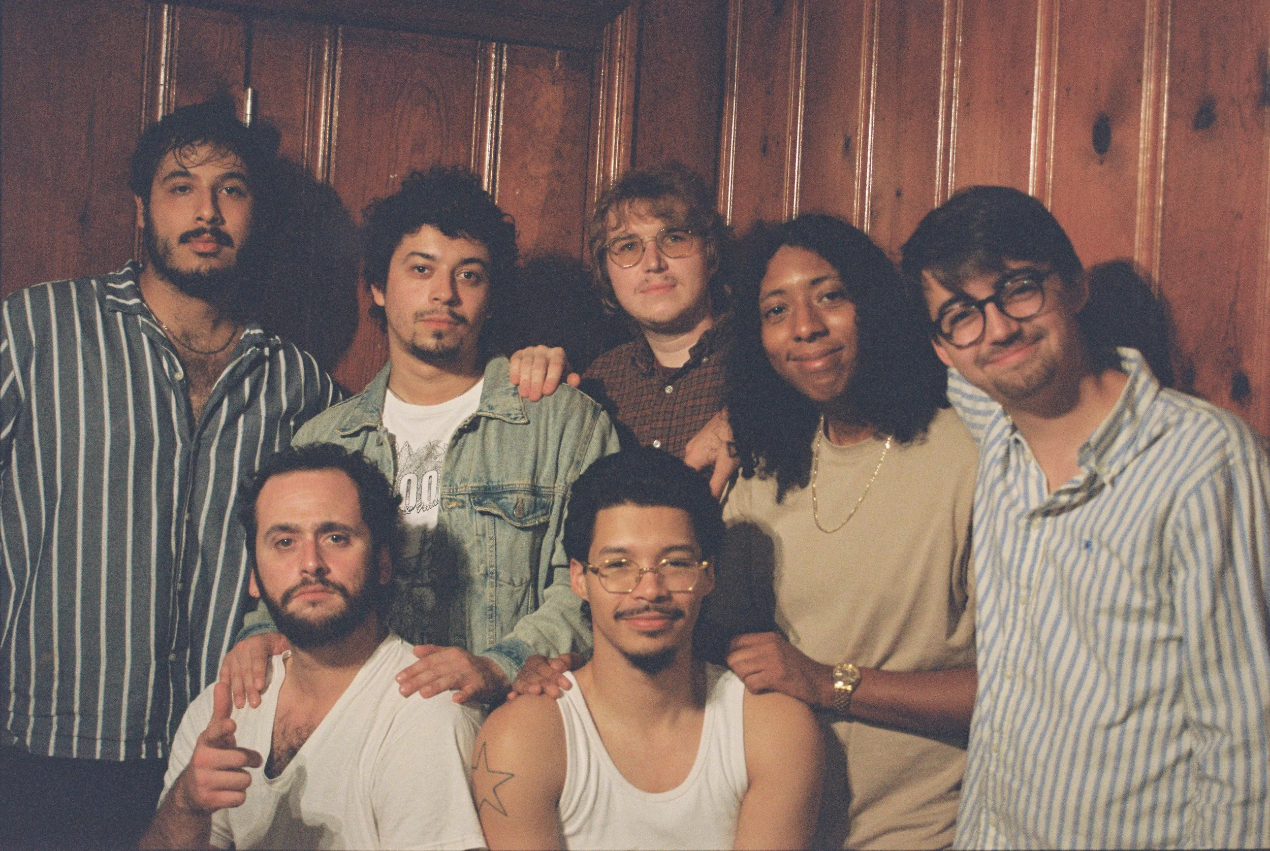 Group of six people posing together in front of wooden wall, some smiling and some with serious expressions, wearing casual clothing including striped shirts, denim jacket, and glasses.