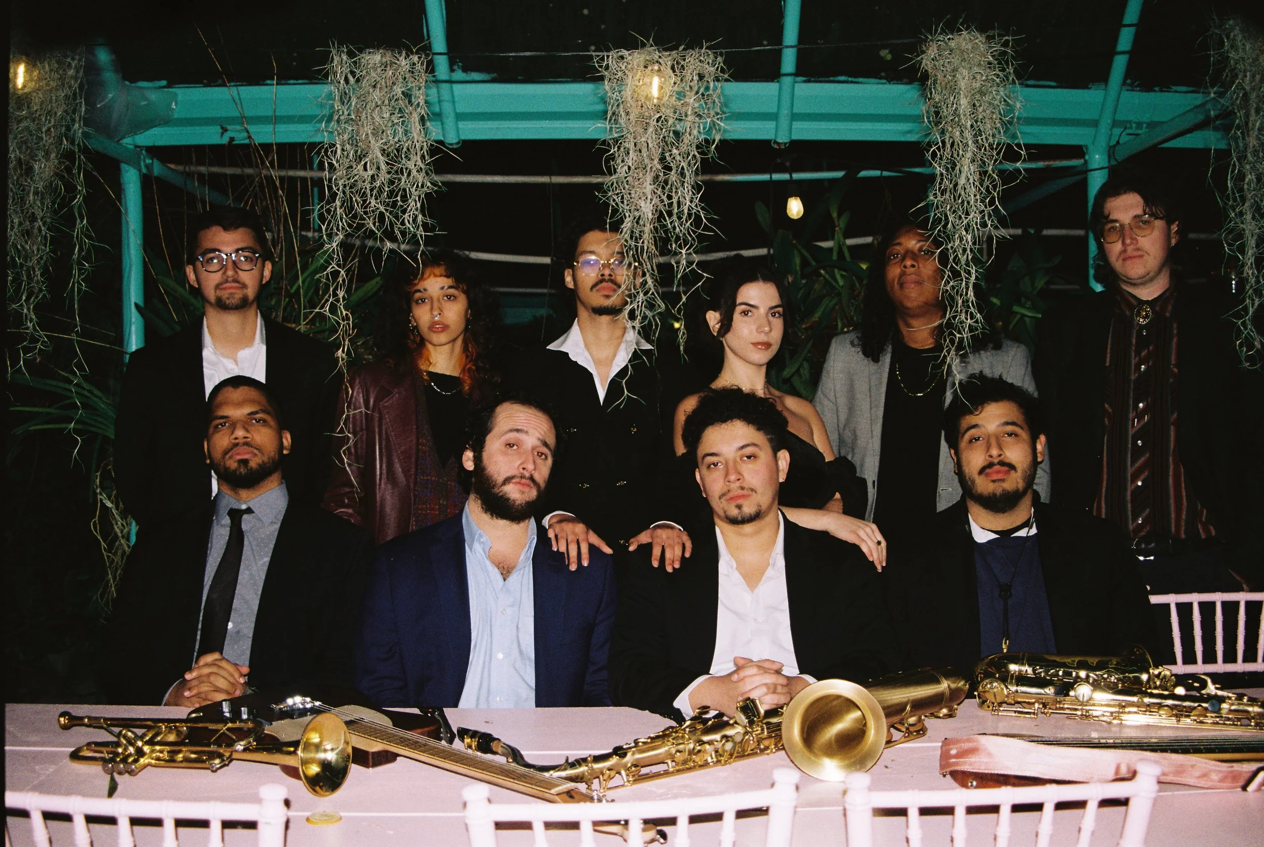 A group of 11 people posing behind a white table with musical instruments, including saxophones, in an indoor or covered outdoor setting with hanging plants and blue lighting.