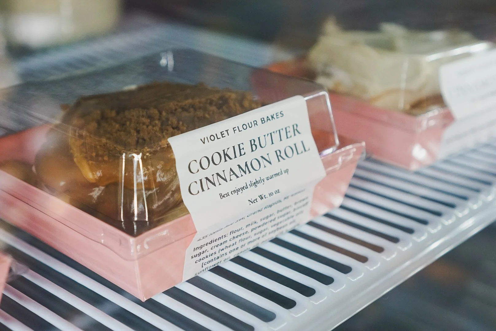 Packet of Violet Flour Baked Cookies with Cookie Butter Cinnamon Roll in a bakery display case.