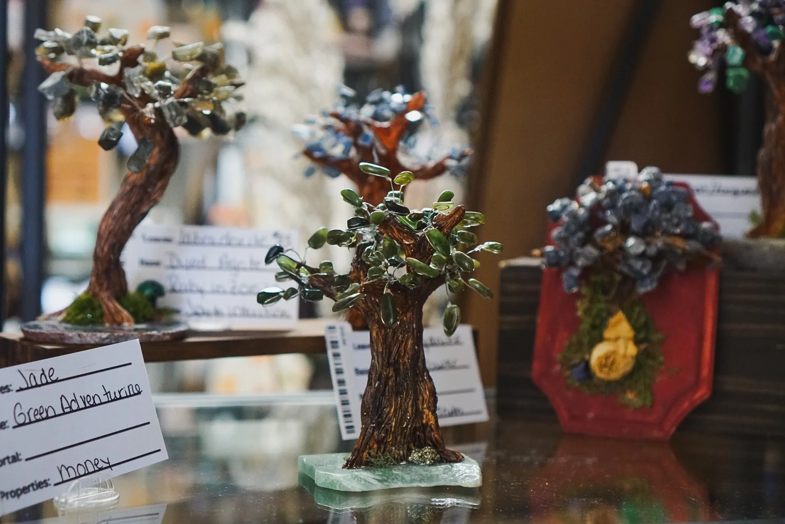 A small, decorative gemstone tree with a brown, textured trunk and dark green, oval-shaped stones for leaves, placed on a marble base, displayed among other gemstone trees and colorful flower arrangements.