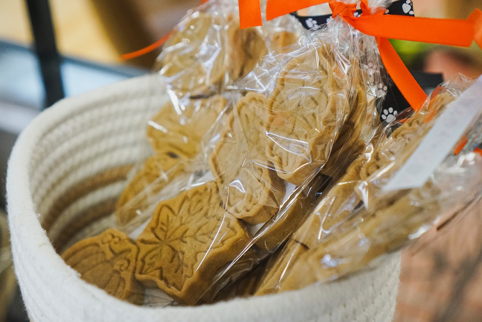 Cookies shaped like leaves and trees in clear plastic bags with orange ribbons inside a white basket.