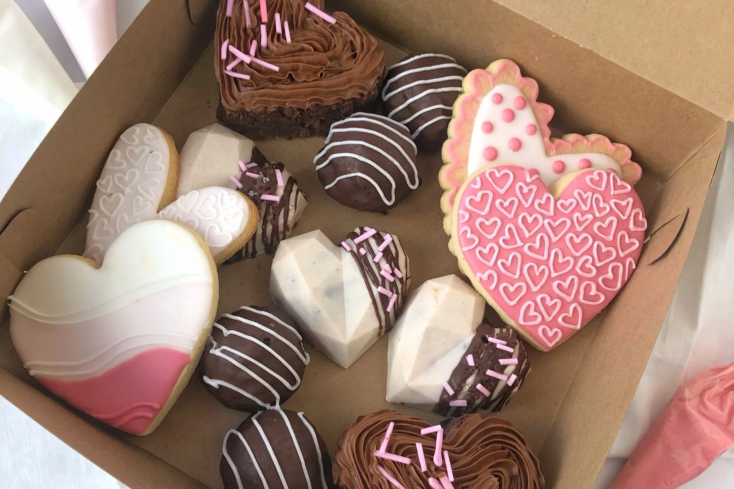 A brown cardboard box containing an assortment of decorated cookies and cupcakes. The cookies are heart-shaped and pink with white hearts, and other assorted shapes with various pink and white icing decorations. The cupcakes are topped with chocolate and pink icing with sprinkles.