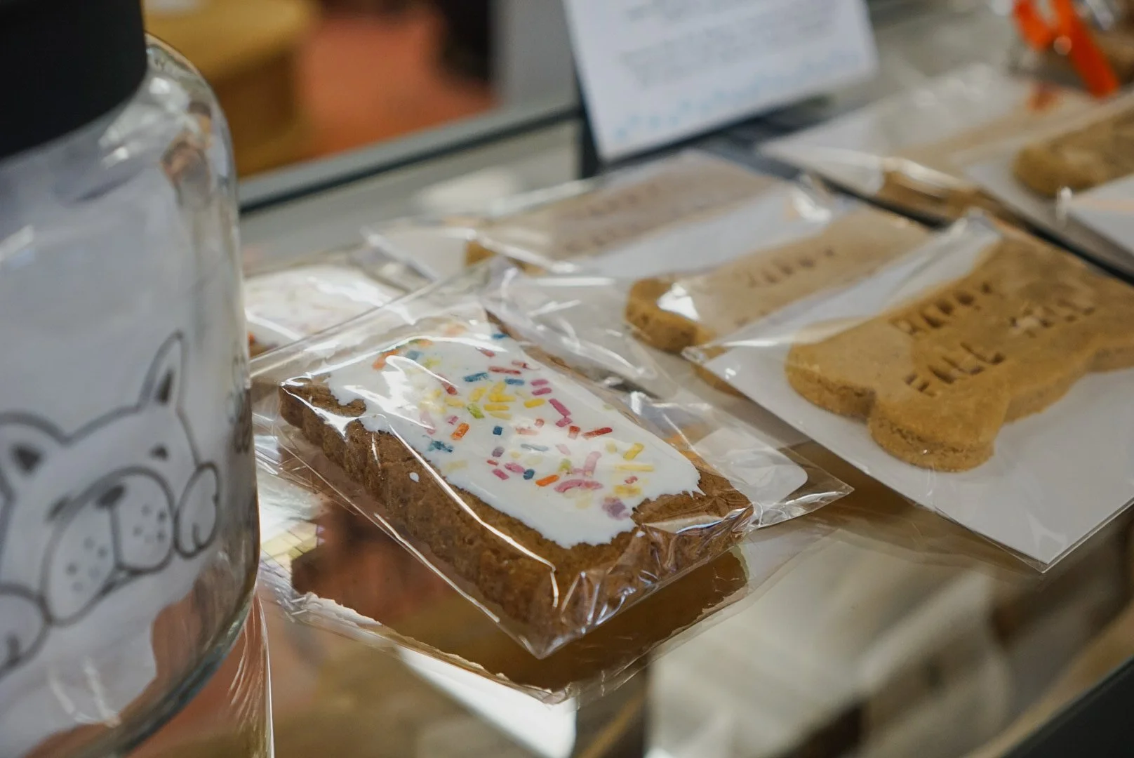 Close-up of decorated cookies shaped like flowers, leaves, and a bunny, arranged in a white box with purple shredded paper filling, on a yellow background with blue and orange flowers and bees.