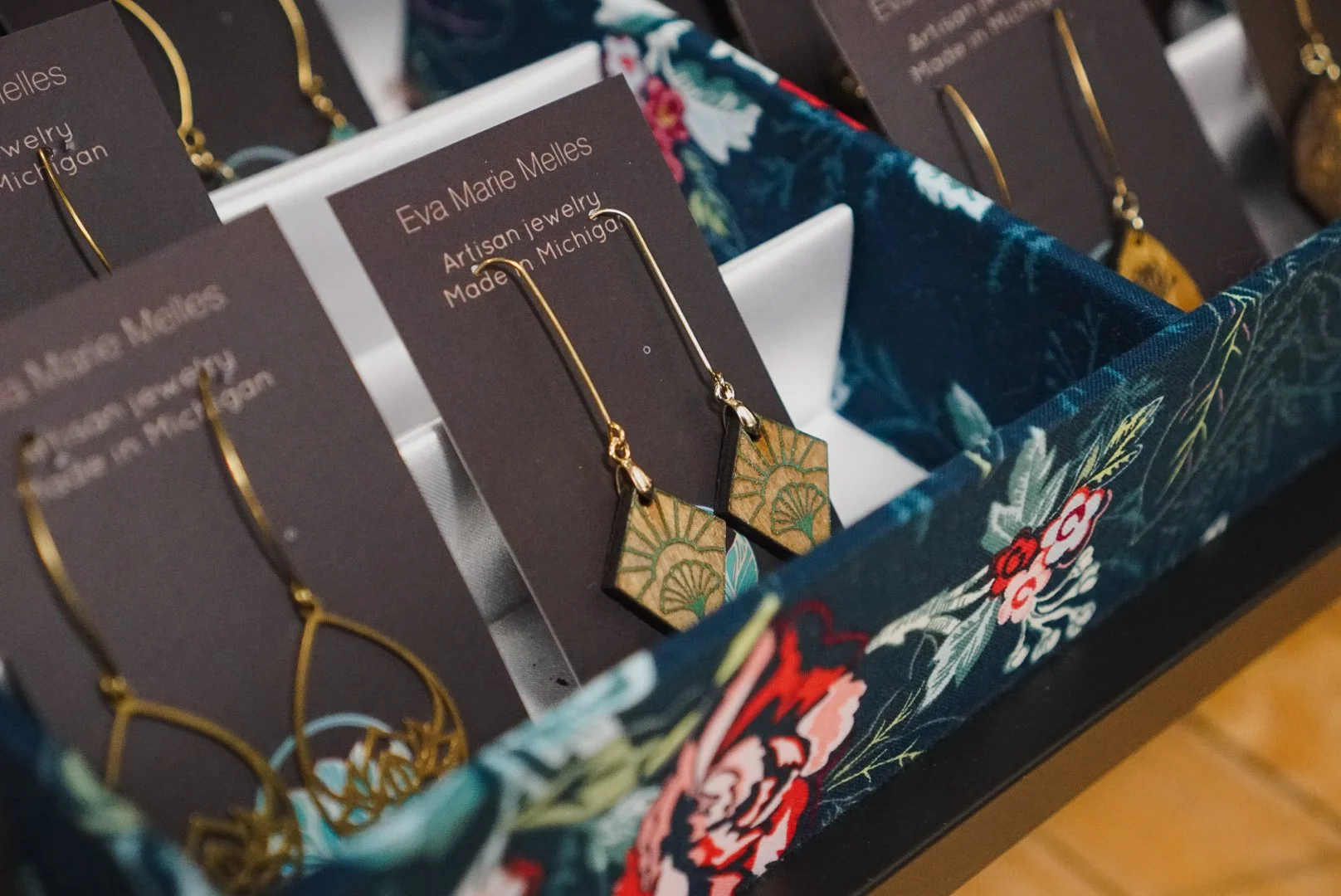 Display of gold and patterned earrings by Eva Marie Melles, artisan jewelry made in Michigan, in a decorated floral fabric-lined box.