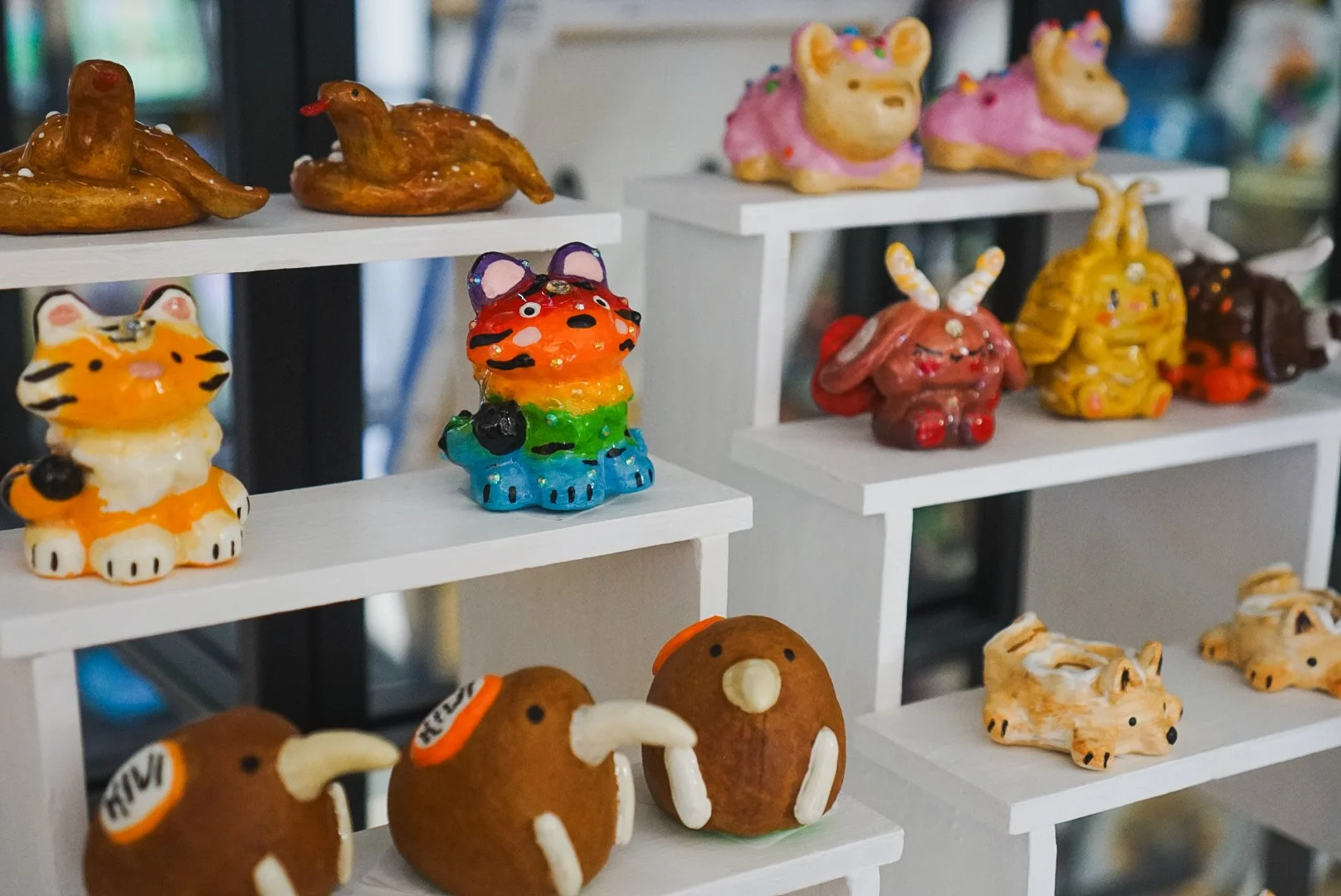 Colorful ceramic figurines of tiger characters, some with cheek blushes and bells, are displayed on a wooden shelf. There are small signs, handwritten notes, and a large illustration of a cute tiger face in the background.