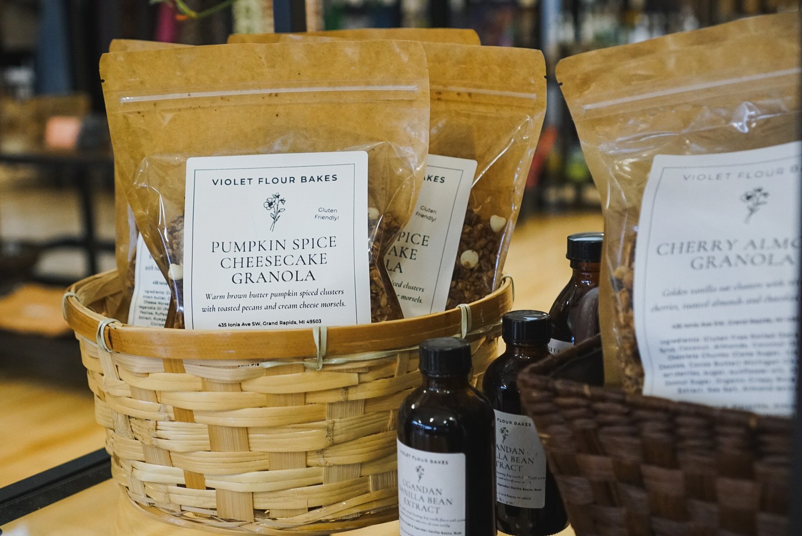 Baskets of packaged granola in a store, including pumpkin spice cheesecake granola and cherry almond granola with labels and small bottles of flavor extract nearby.