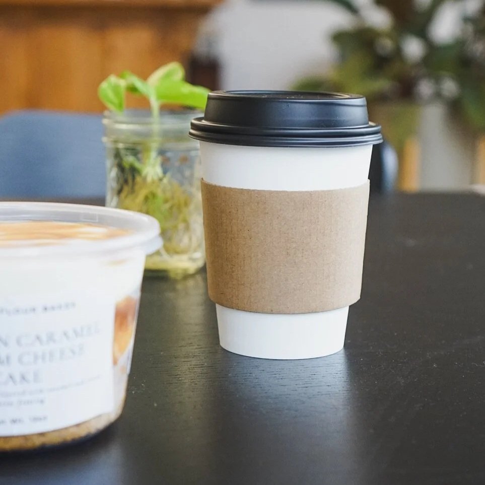 A white disposable coffee cup with a black lid and a brown cardboard sleeve on black table, with a blurred container of food and a small plant in the background.