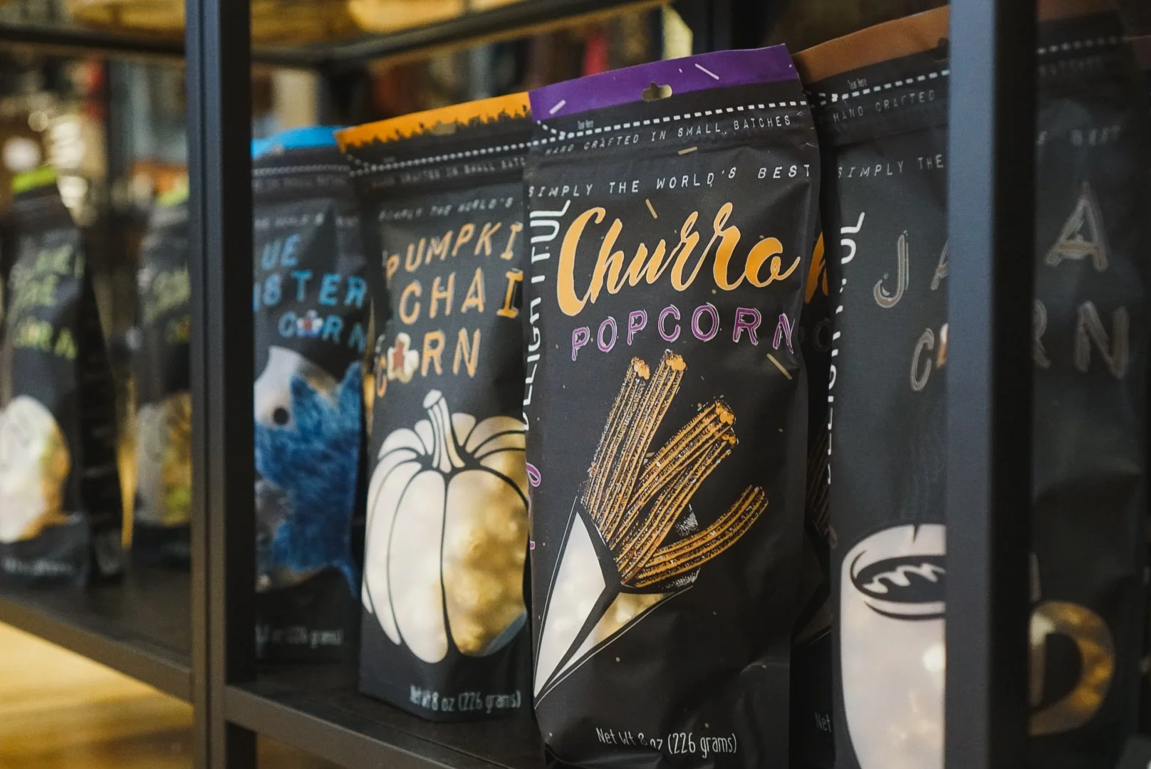 Black snack bag labeled "Churchill Popcorn" with an illustration of caramel-coated popcorn sticks, placed on a store shelf with other snack bags behind.