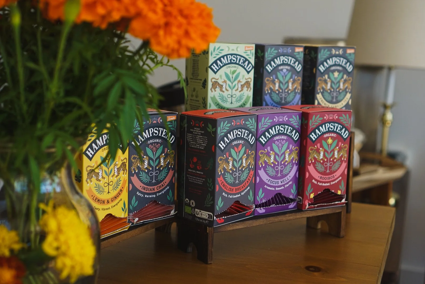 Assorted packs of Hampstead organic tea boxes on a wooden shelf, with a blurred bouquet of oranges, yellow, and green flowers in the foreground.
