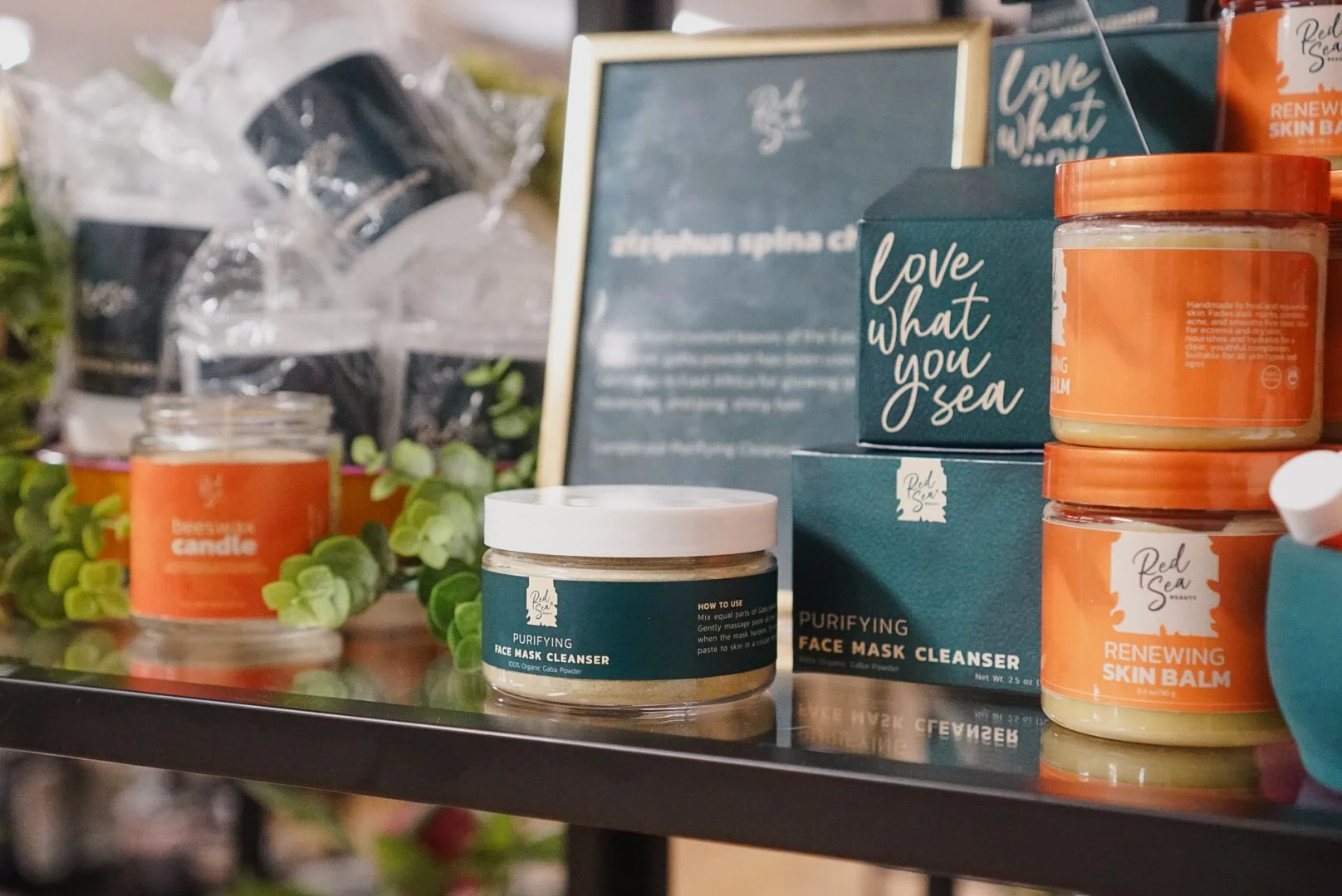 Display of skincare products including face mask cleansers and skin balms with a chalkboard and decorative greenery in the background.