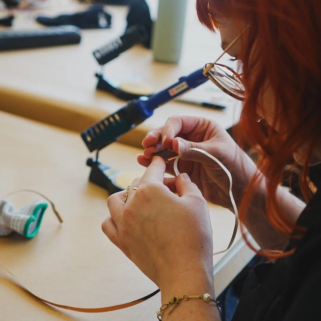 Person with red hair and glasses working with soldering equipment on a small electronic component at a workbench with tools and materials in the background.