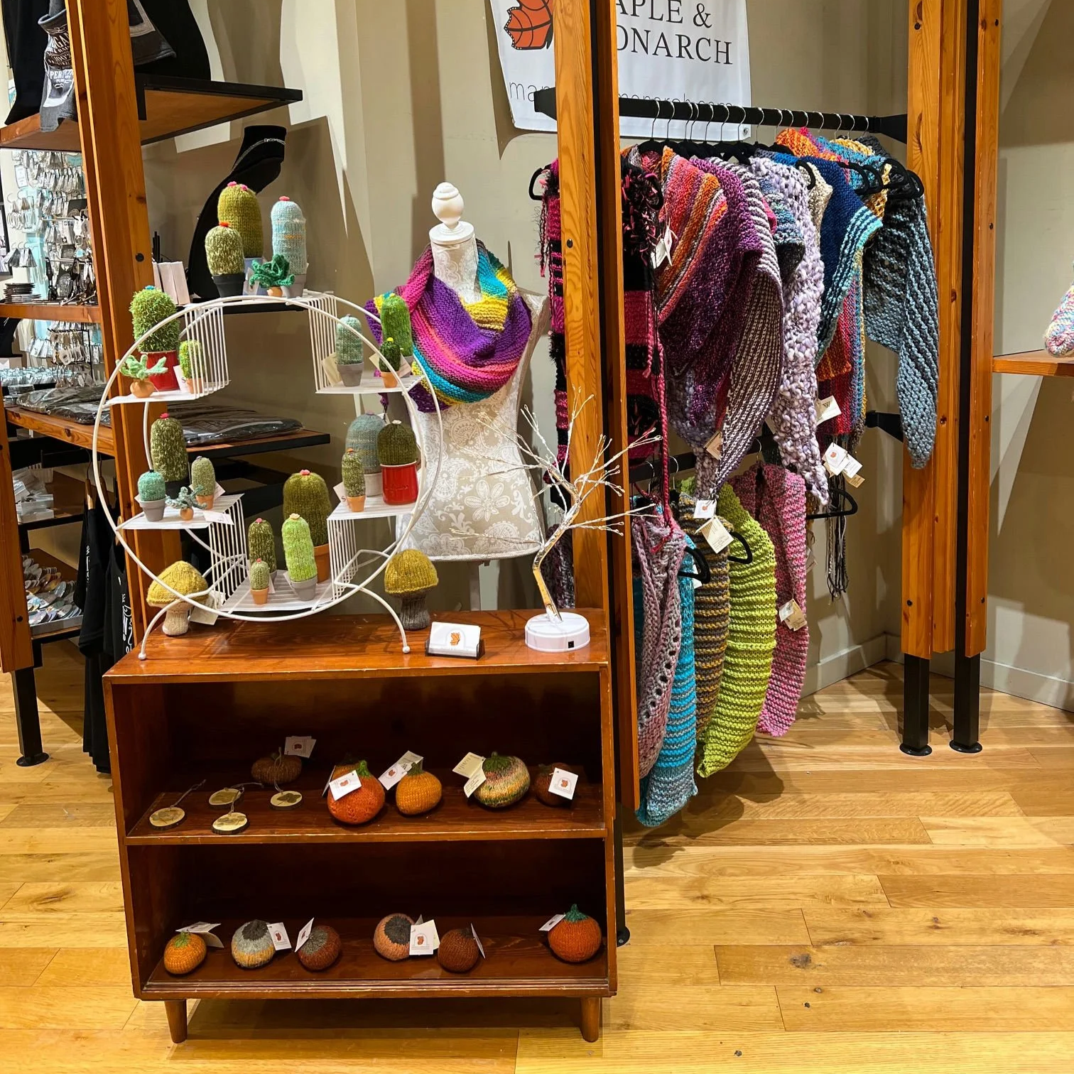 Display of colorful knitted accessories, clothing, and decorations in a store, including scarves, sweaters, hats, and cactus-shaped knitted pouches.