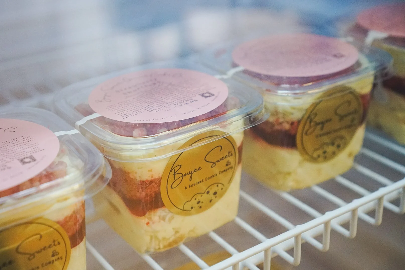 Plastic containers of layered dessert in a refrigerated display, labeled with "Boujee Sweets" and featuring a pink round top and a yellow base layered with cake and cream.