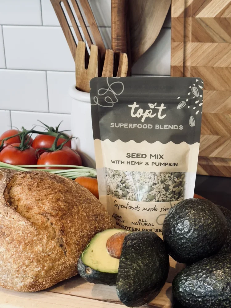 A loaf of bread, a halved avocado, three whole avocados, a bag of seed mix with hemp and pumpkin, a bunch of cherry tomatoes, and some wooden kitchen utensils on a countertop.
